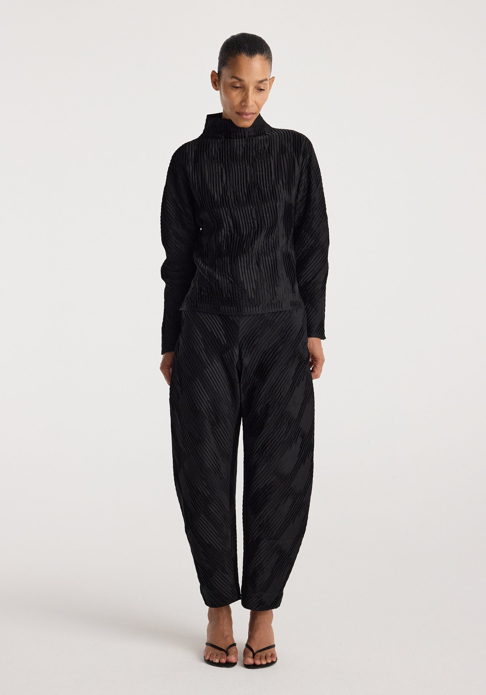 Woman wearing black Plissé satin trousers | black with a matching long-sleeve top, standing against a plain light background and looking downward.