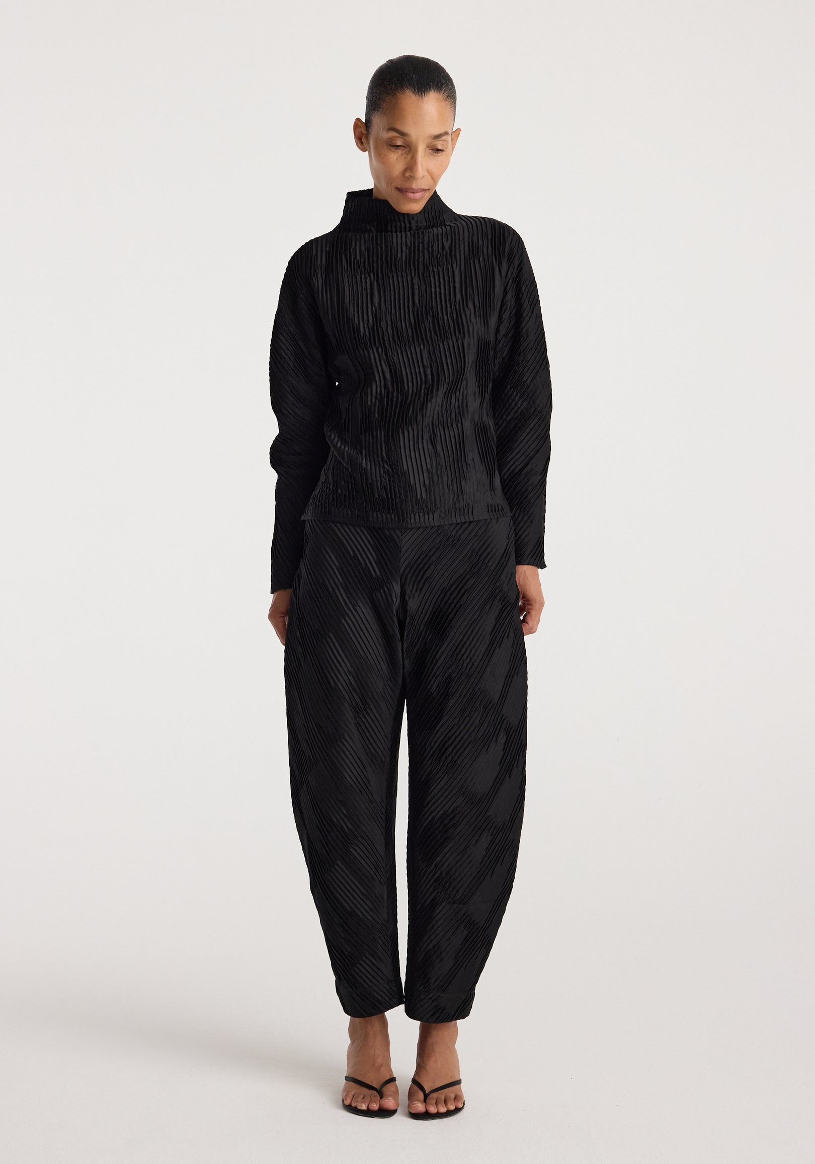 Woman wearing black Plissé satin trousers | black with a matching long-sleeve top, standing against a plain light background and looking downward.