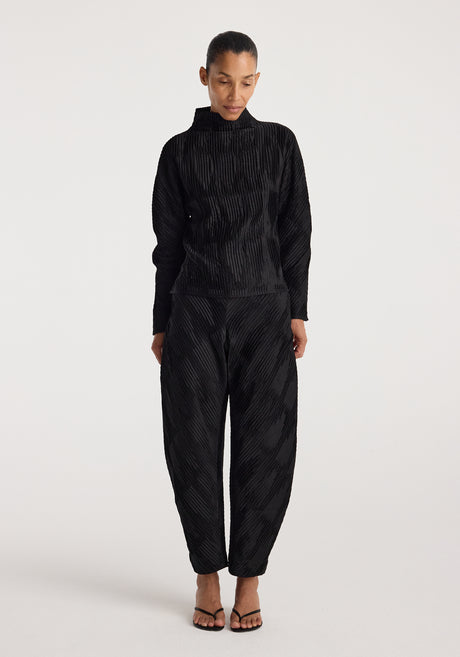 Woman wearing black Plissé satin trousers | black with a matching long-sleeve top, standing against a plain light background and looking downward.