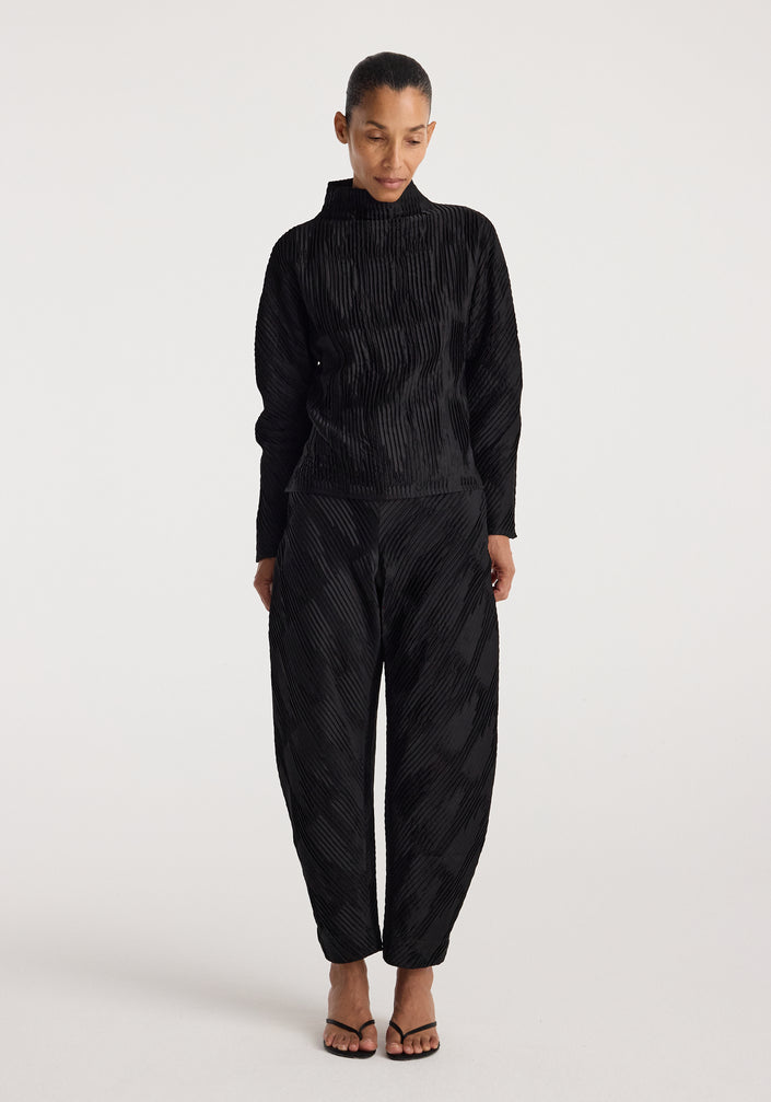 Woman wearing black Plissé satin trousers | black with a matching long-sleeve top, standing against a plain light background and looking downward.
