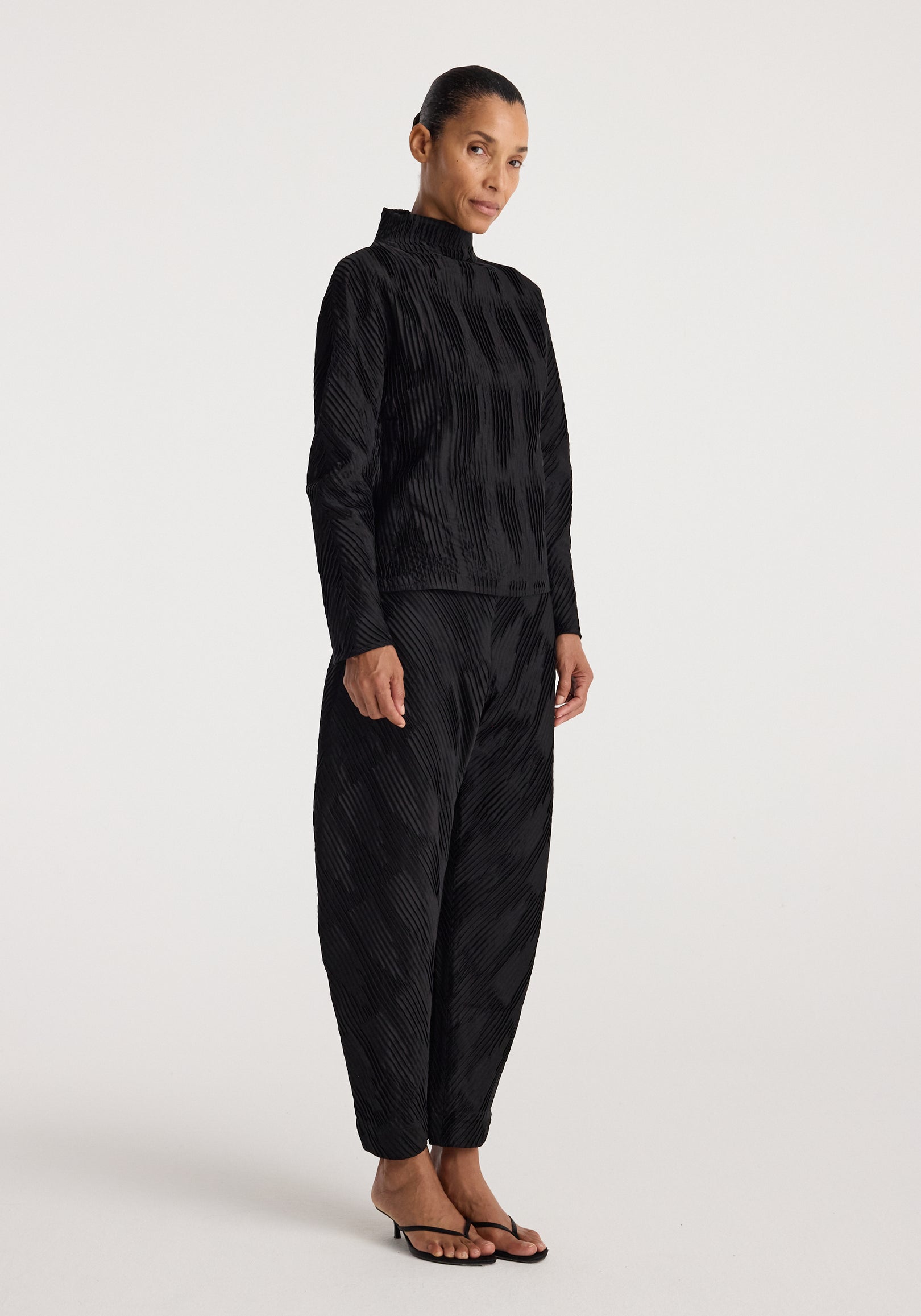 Woman wearing black textured long-sleeve top paired with plissé satin trousers | black and black heeled sandals against a plain light background.