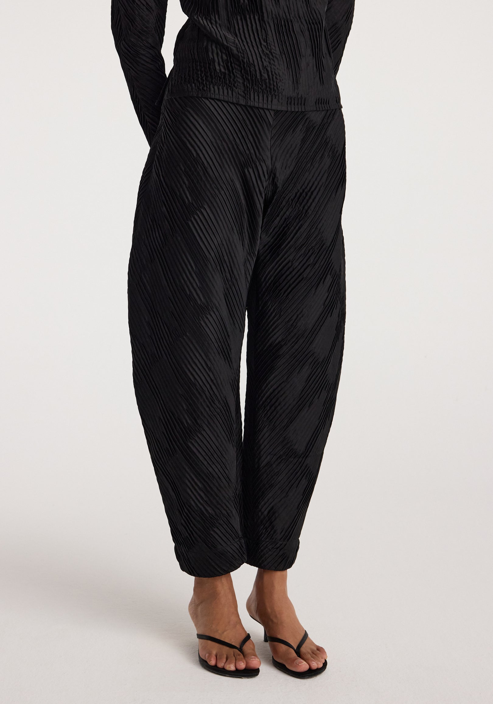 Lower body view of a person wearing black Plissé satin trousers | black with a textured pleated pattern, paired with black strappy heeled sandals on a neutral background.