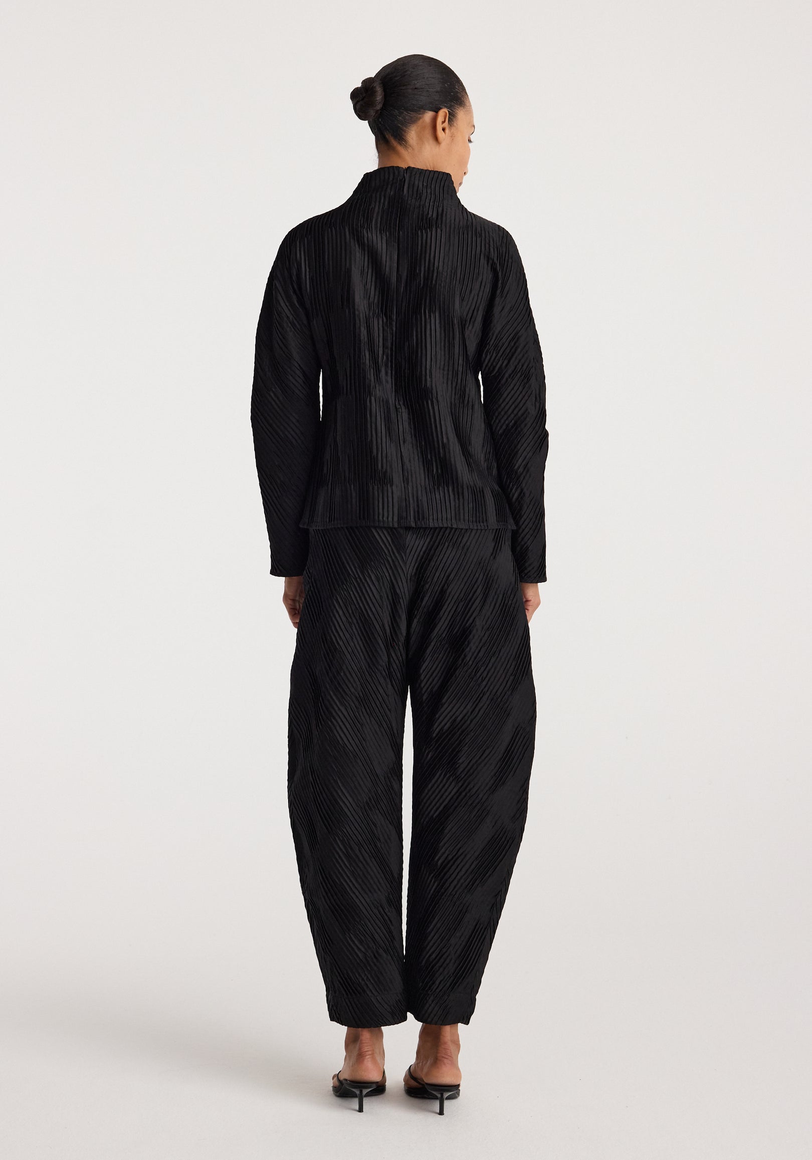 Woman seen from the back wearing black Plissé satin trousers | black with a matching long-sleeve top and black heeled sandals on a plain light background.