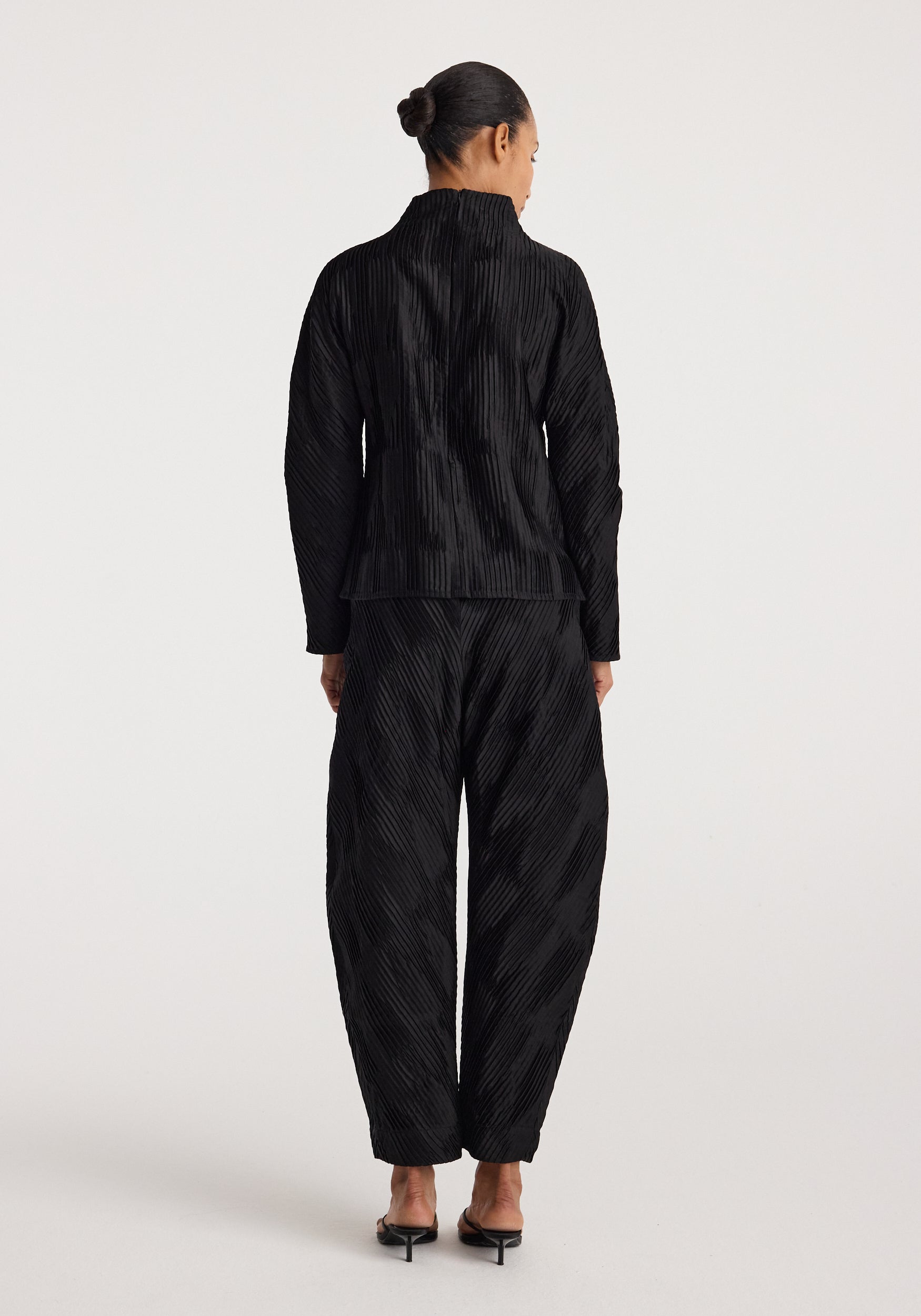 Woman seen from the back wearing black Plissé satin trousers | black with a matching long-sleeve top and black heeled sandals on a plain light background.