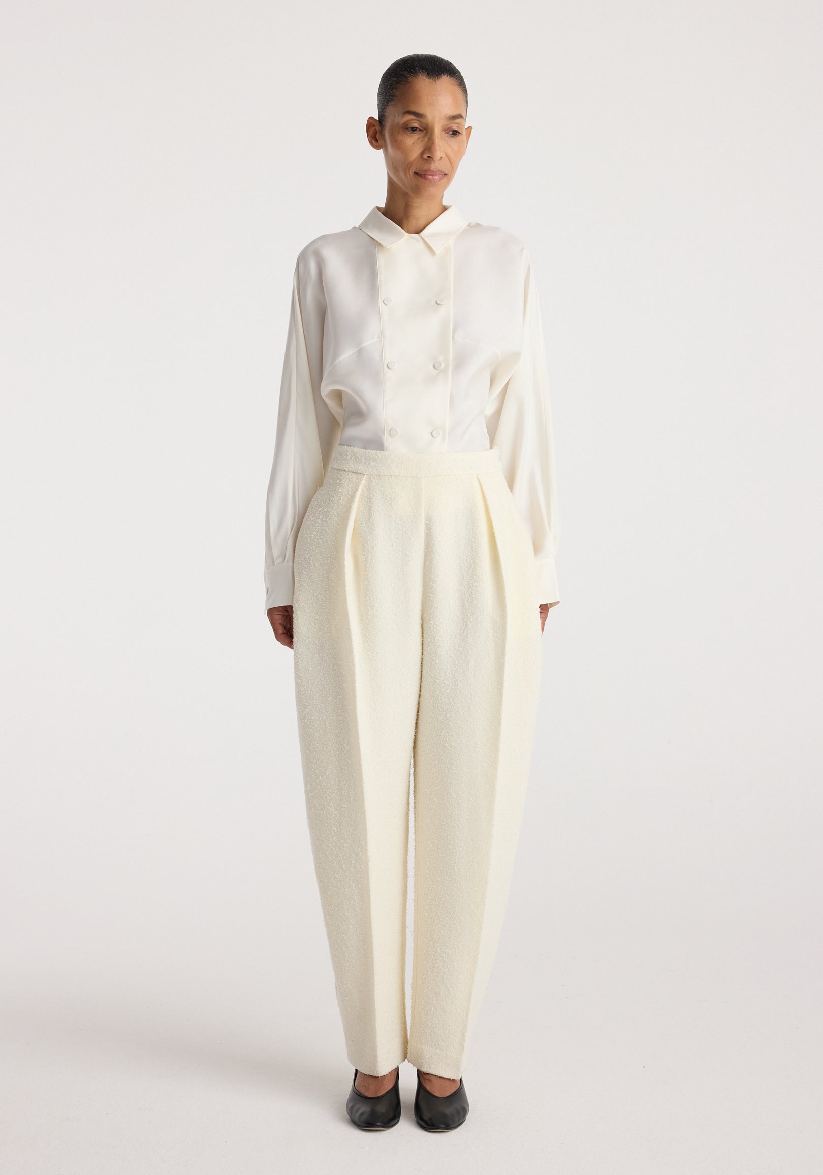 Woman wearing Puck - Sculptural bouclé tailored trousers | cream with a white long-sleeve buttoned shirt and black shoes, standing against a plain white background.
