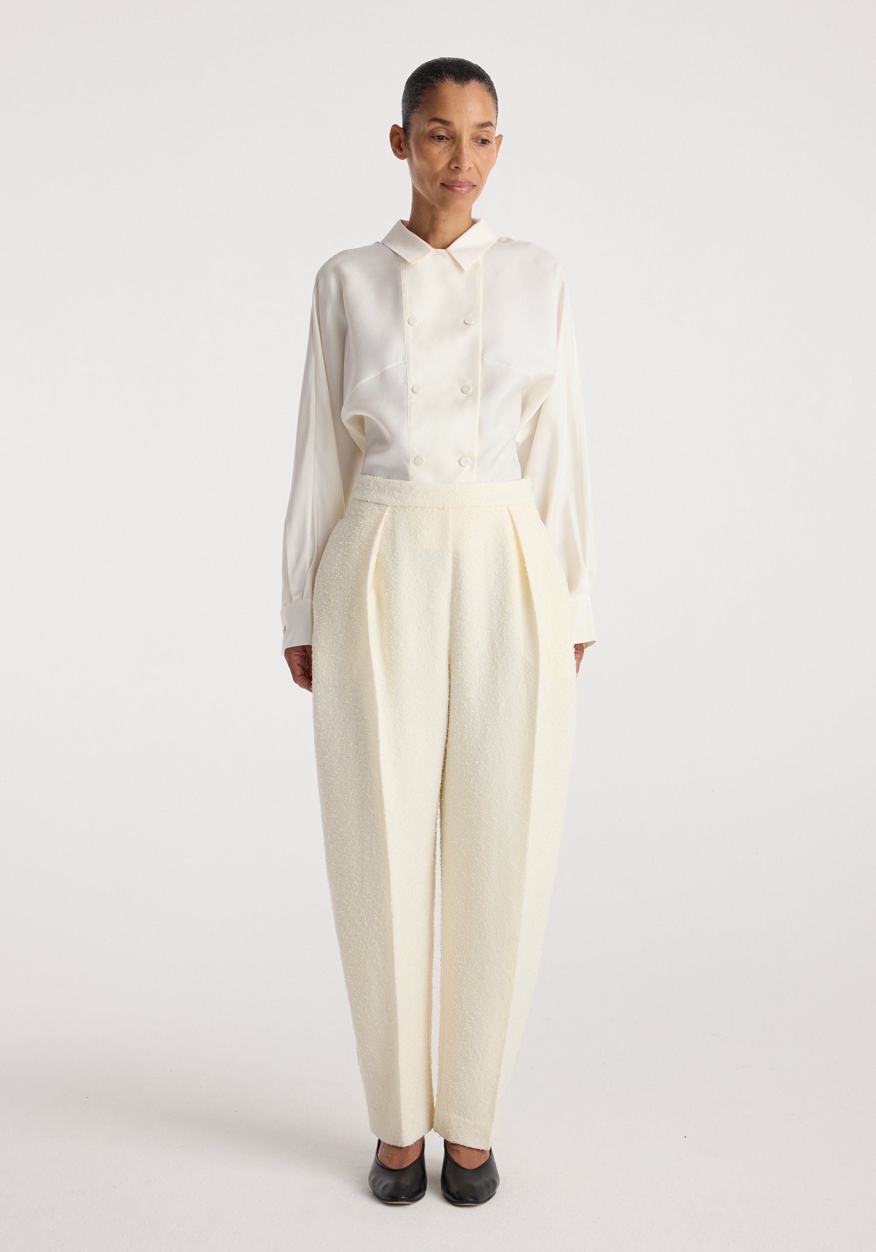 Woman wearing Puck - Sculptural bouclé tailored trousers | cream with a white long-sleeve buttoned shirt and black shoes, standing against a plain white background.