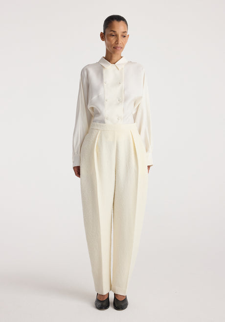 Woman wearing Puck - Sculptural bouclé tailored trousers | cream with a white long-sleeve buttoned shirt and black shoes, standing against a plain white background.