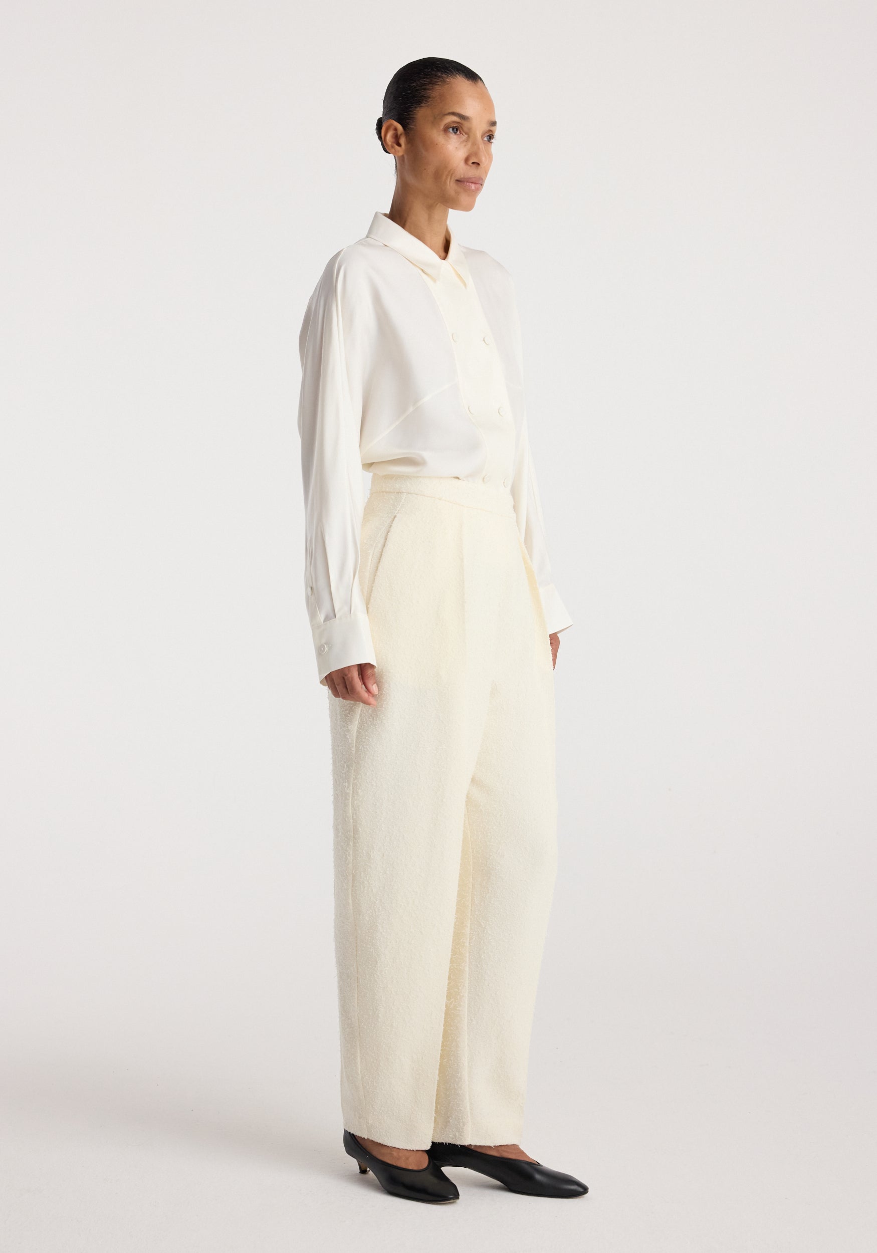 Woman wearing Puck - Sculptural bouclé tailored trousers | cream paired with a white long-sleeve collared shirt and black low-heel shoes against a plain background.