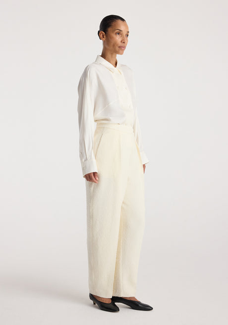 Woman wearing Puck - Sculptural bouclé tailored trousers | cream with a white long-sleeve buttoned shirt and black shoes, standing against a plain white background.
