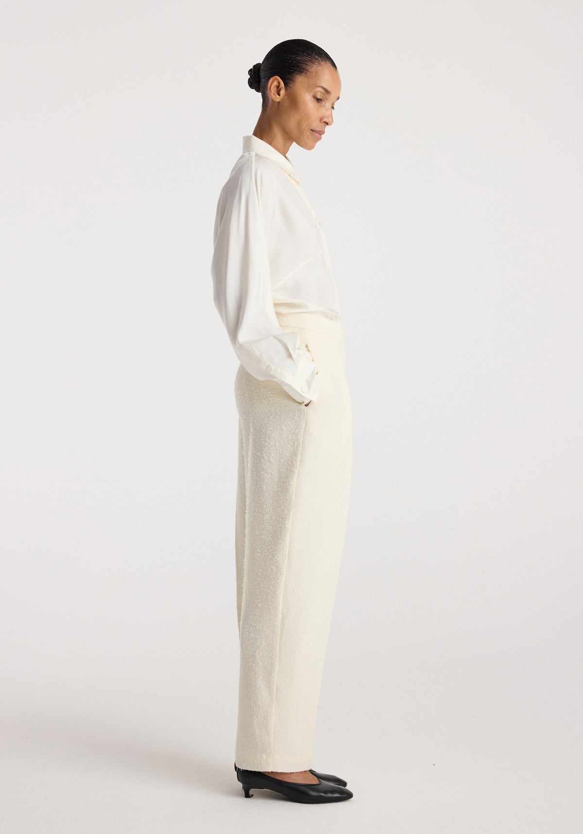 Woman wearing cream-colored Puck - Sculptural bouclé tailored trousers | cream paired with a white long-sleeve blouse and black heels, standing in profile against a plain background.