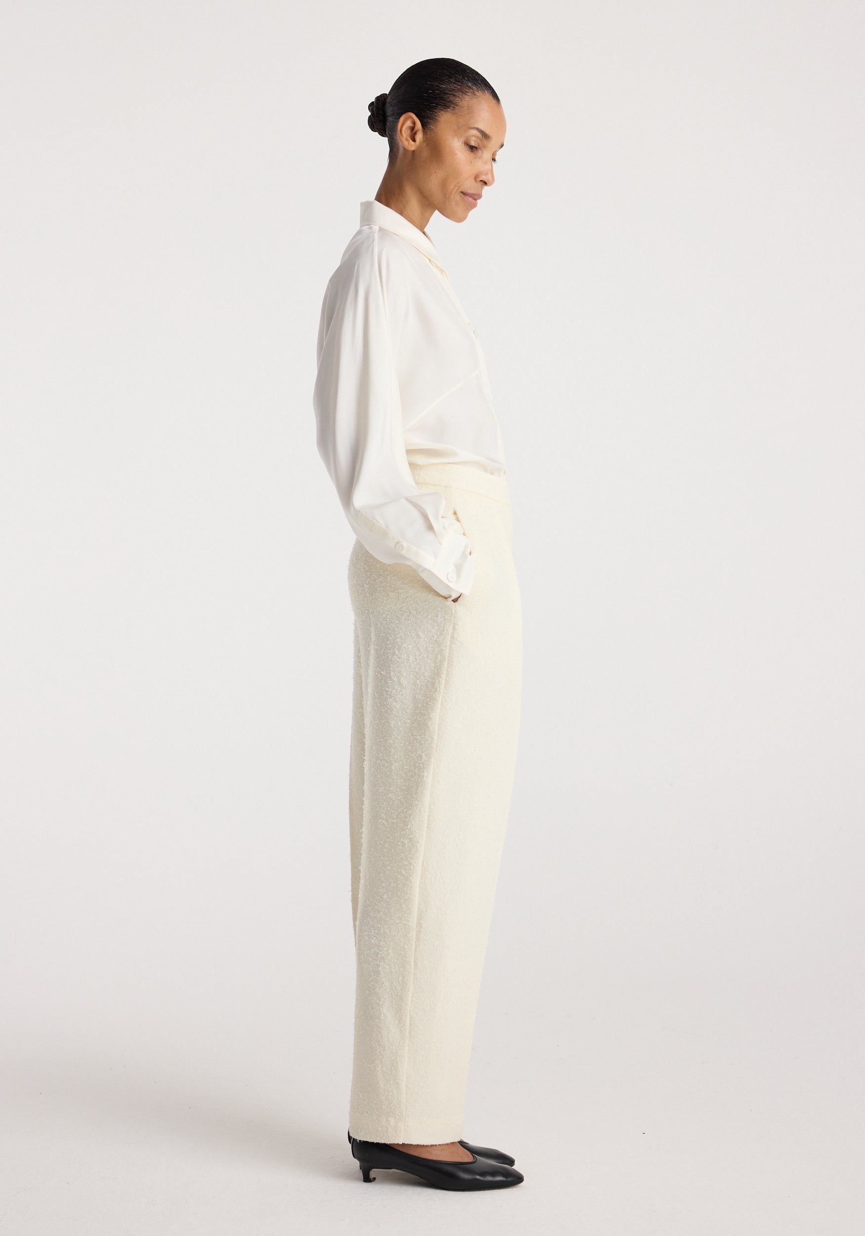 Woman wearing cream-colored Puck - Sculptural bouclé tailored trousers | cream paired with a white long-sleeve blouse and black heels, standing in profile against a plain background.