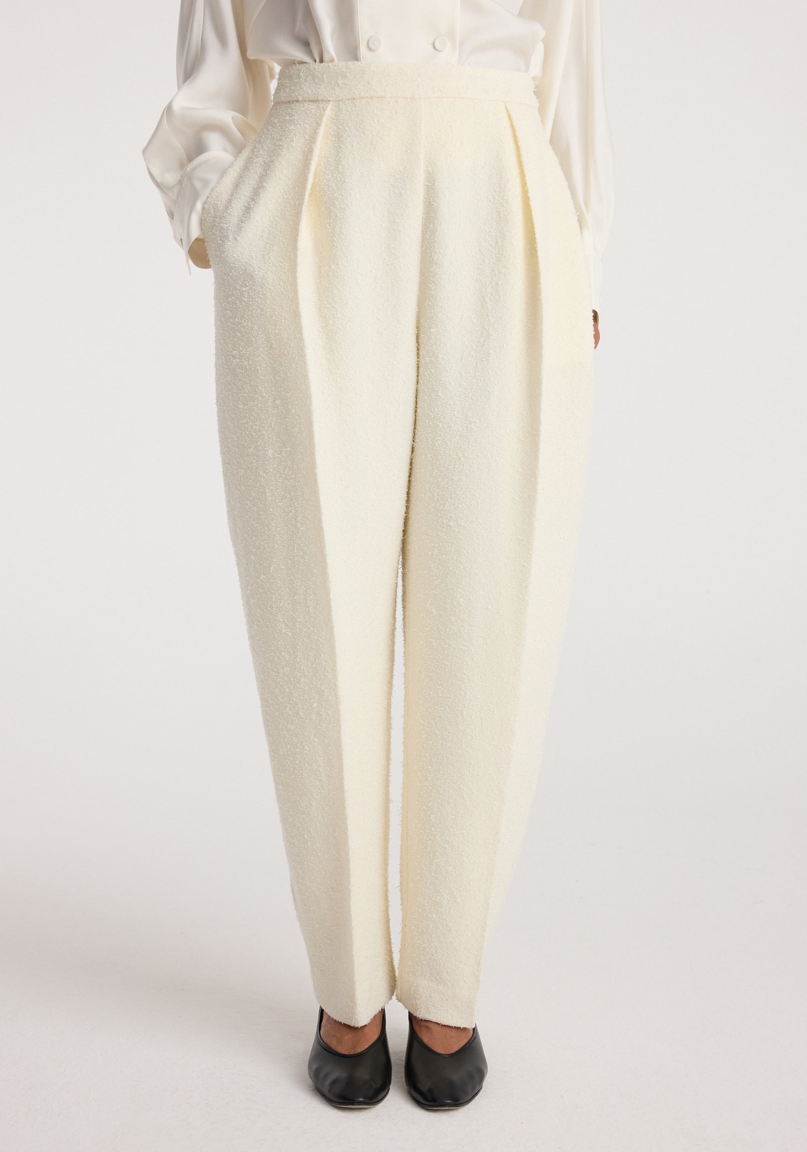Front view of a model wearing Puck - Sculptural bouclé tailored trousers | cream, styled with a white blouse and black shoes, showcasing the textured fabric and pleated design.