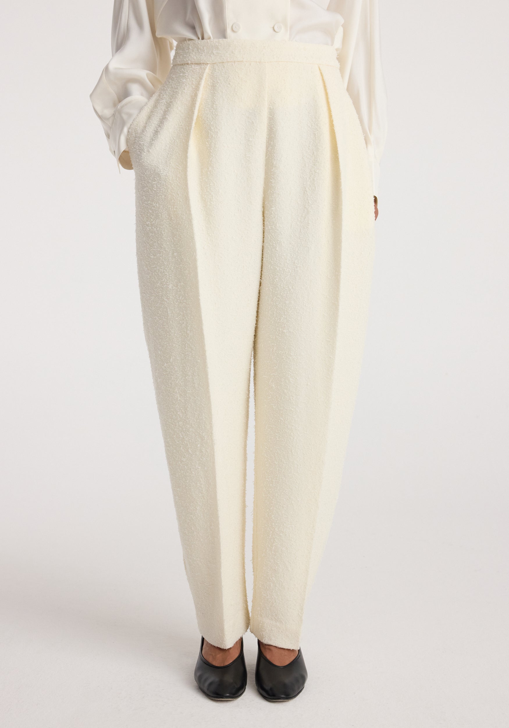 Front view of a model wearing Puck - Sculptural bouclé tailored trousers | cream, styled with a white blouse and black shoes, showcasing the textured fabric and pleated design.