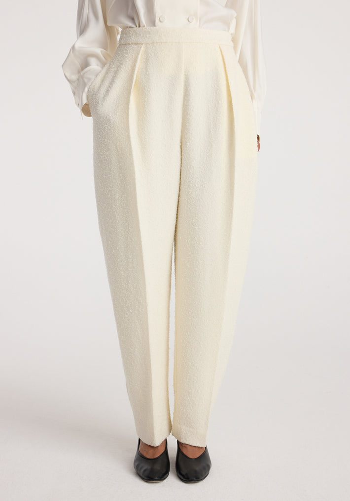 Front view of a model wearing Puck - Sculptural bouclé tailored trousers | cream, styled with a white blouse and black shoes, showcasing the textured fabric and pleated design.
