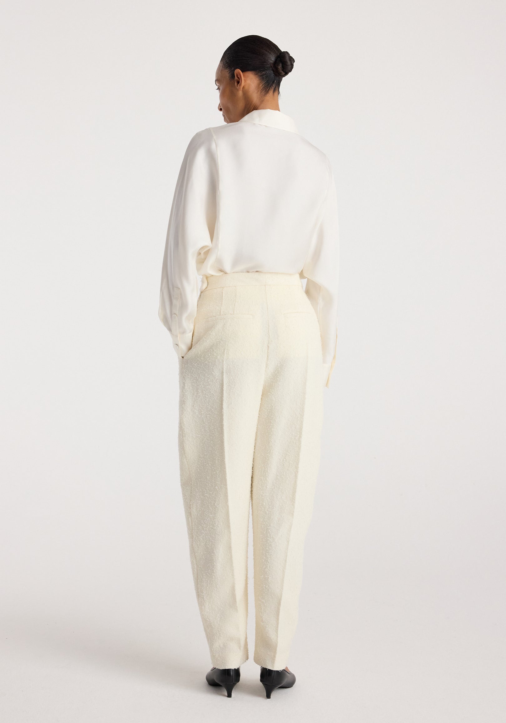 Woman seen from behind wearing Puck - Sculptural bouclé tailored trousers | cream with a tucked-in white blouse and black pointed heels, standing against a plain light background.
