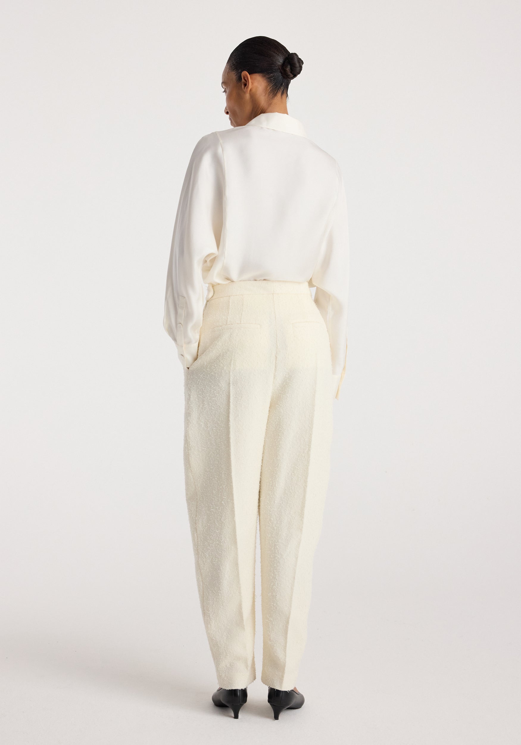 Woman seen from behind wearing Puck - Sculptural bouclé tailored trousers | cream with a tucked-in white blouse and black pointed heels, standing against a plain light background.