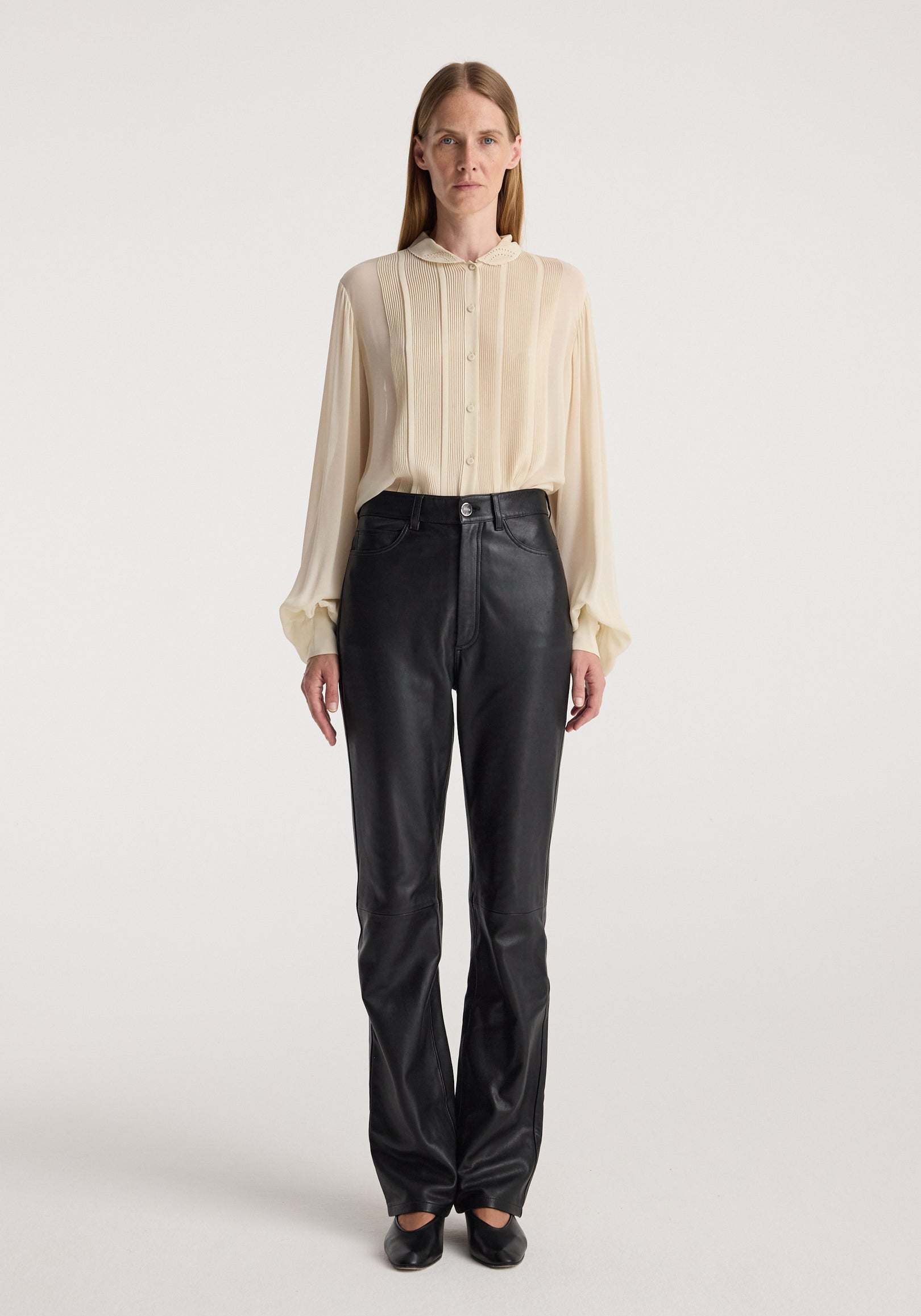 Woman wearing high-waist straight leather trousers | black paired with a light beige long-sleeve blouse and black shoes, standing against a plain background.