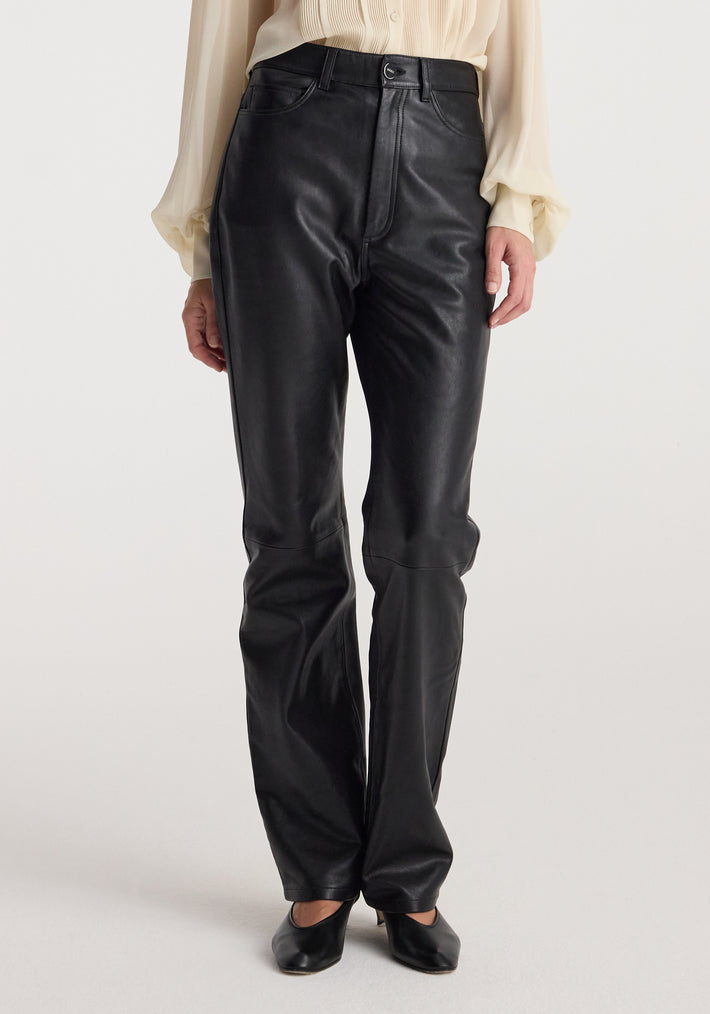 Front view of a person wearing high-waist straight leather trousers | black paired with a light-colored blouse and black shoes against a neutral background.
