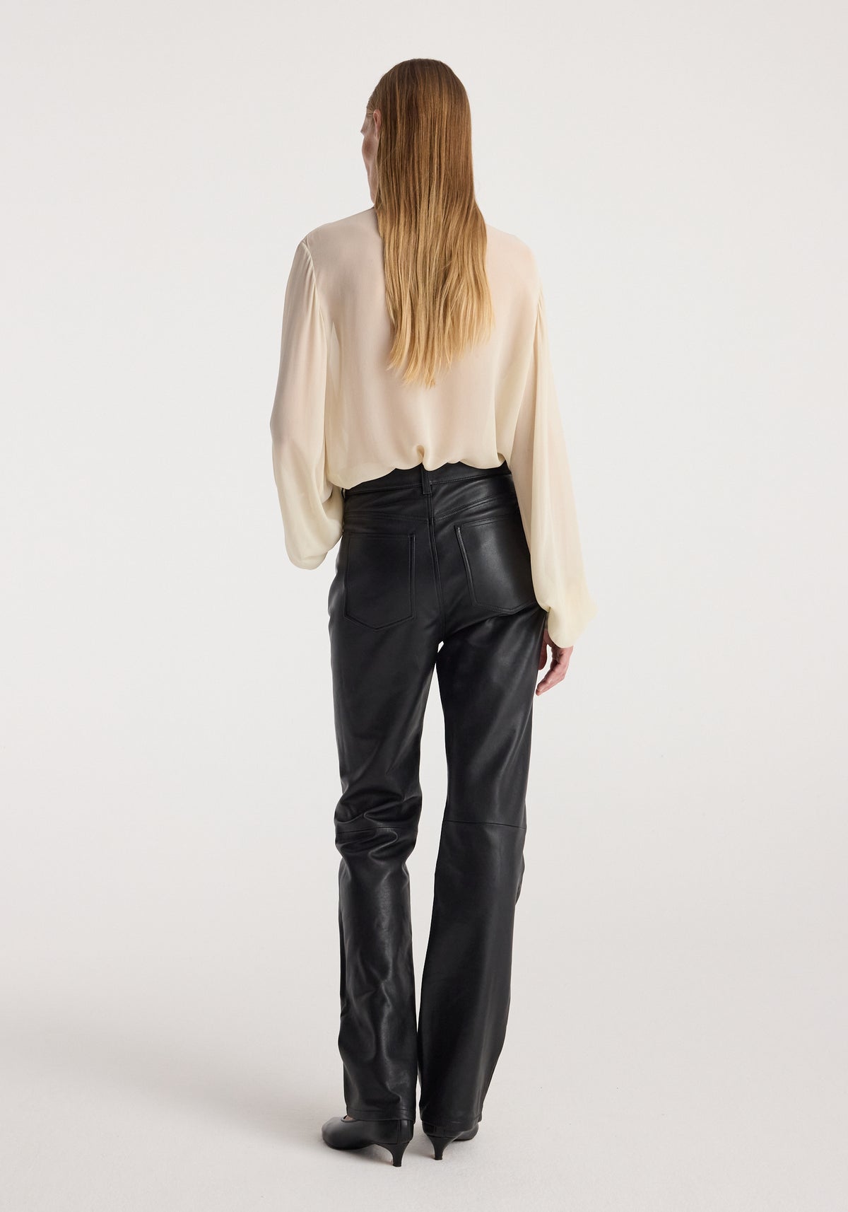 Woman wearing high-waist straight leather trousers | black with a cream-colored long sleeve blouse, shown from the back against a plain light background.