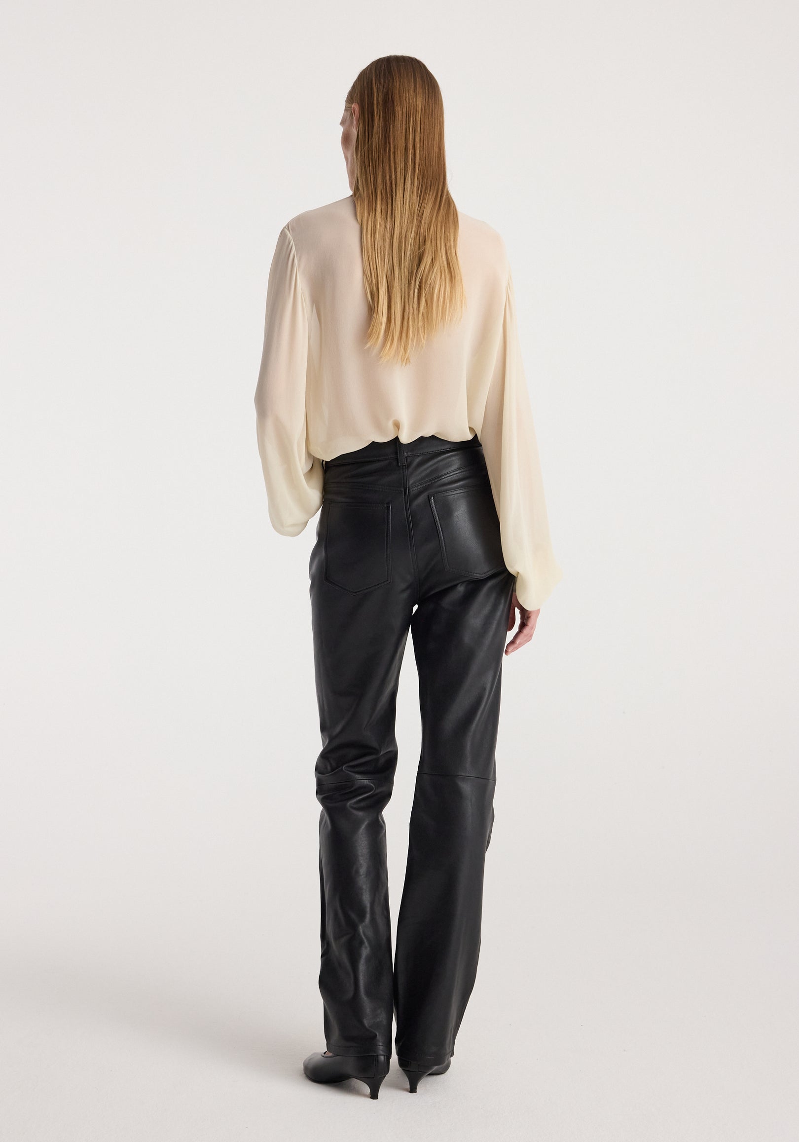 Woman wearing high-waist straight leather trousers | black with a cream-colored long sleeve blouse, shown from the back against a plain light background.