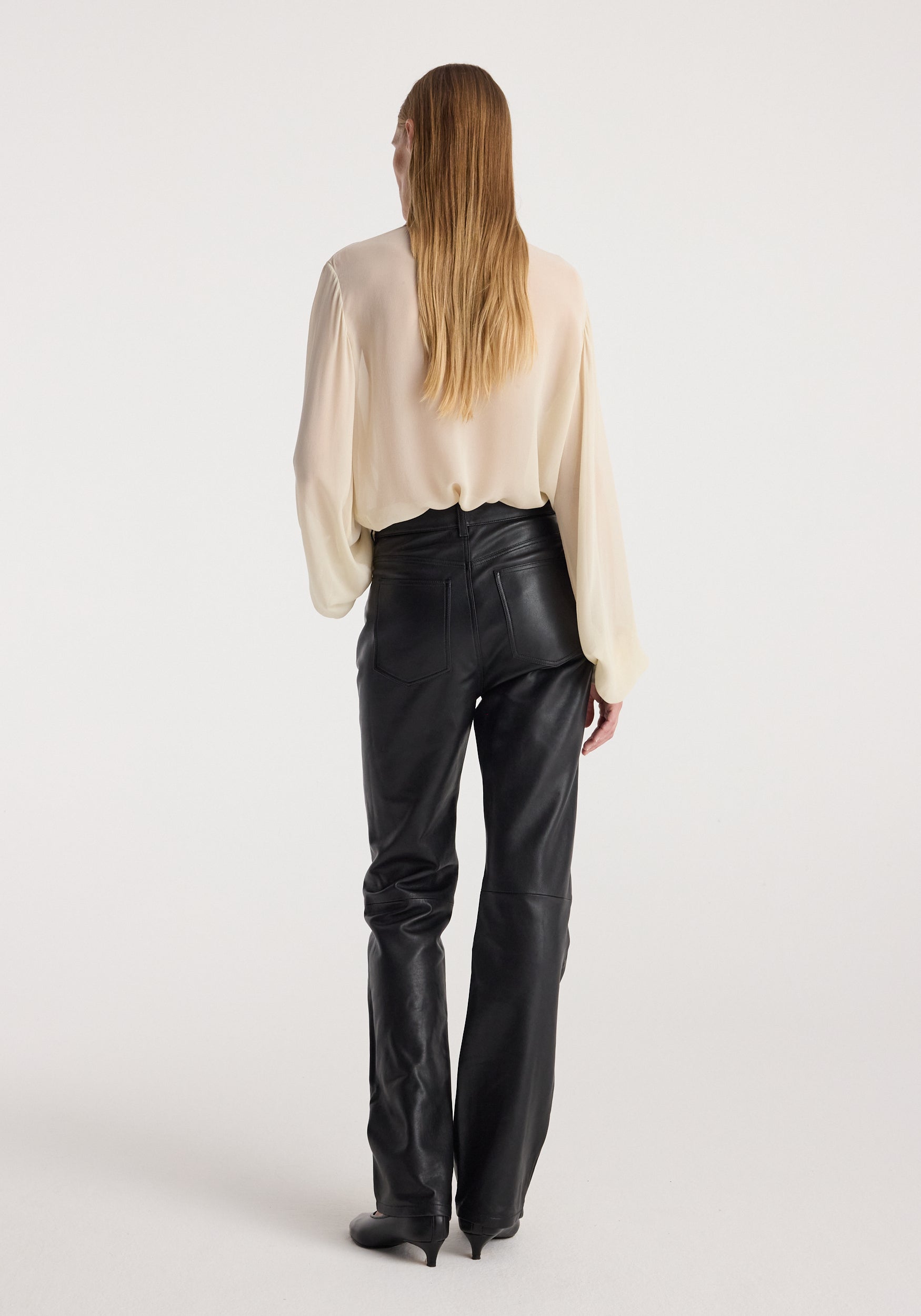 Woman wearing high-waist straight leather trousers | black with a cream-colored long sleeve blouse, shown from the back against a plain light background.