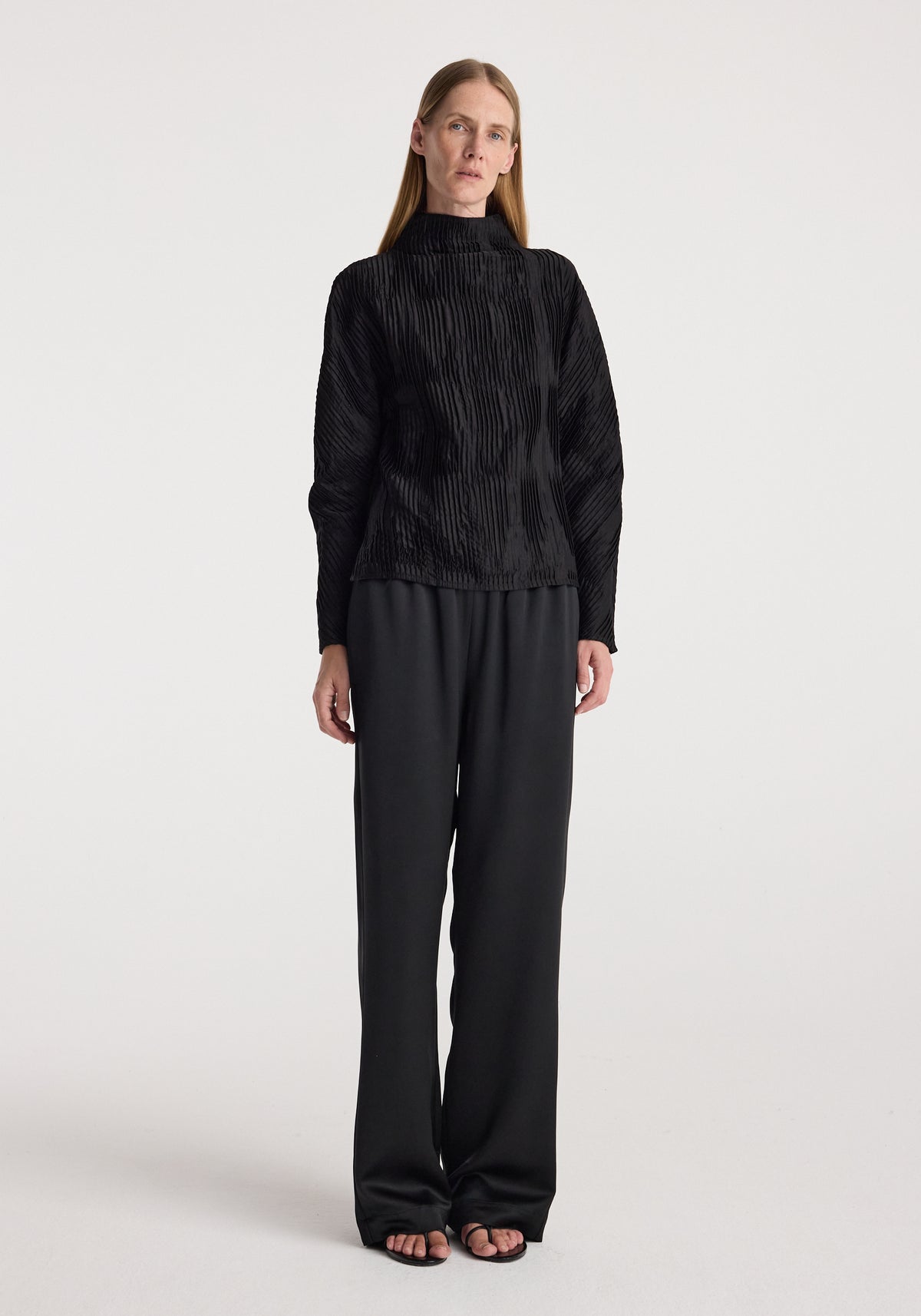 Woman wearing relaxed fit satin trousers | black paired with a textured black long-sleeve top, standing against a plain white background.