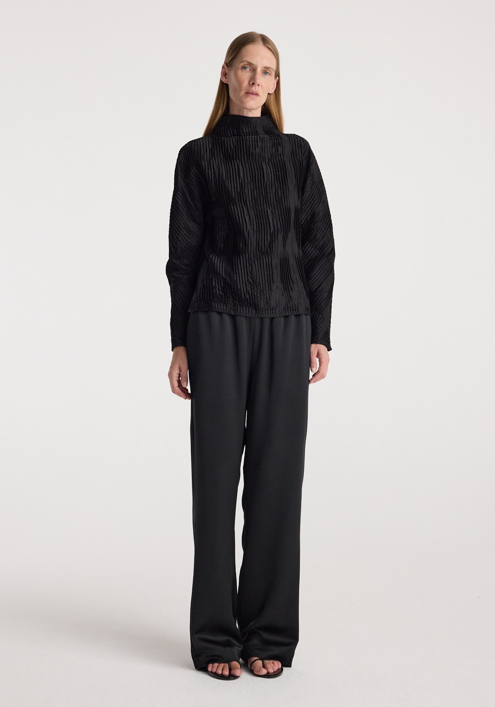 Woman wearing relaxed fit satin trousers | black paired with a textured black long-sleeve top, standing against a plain white background.