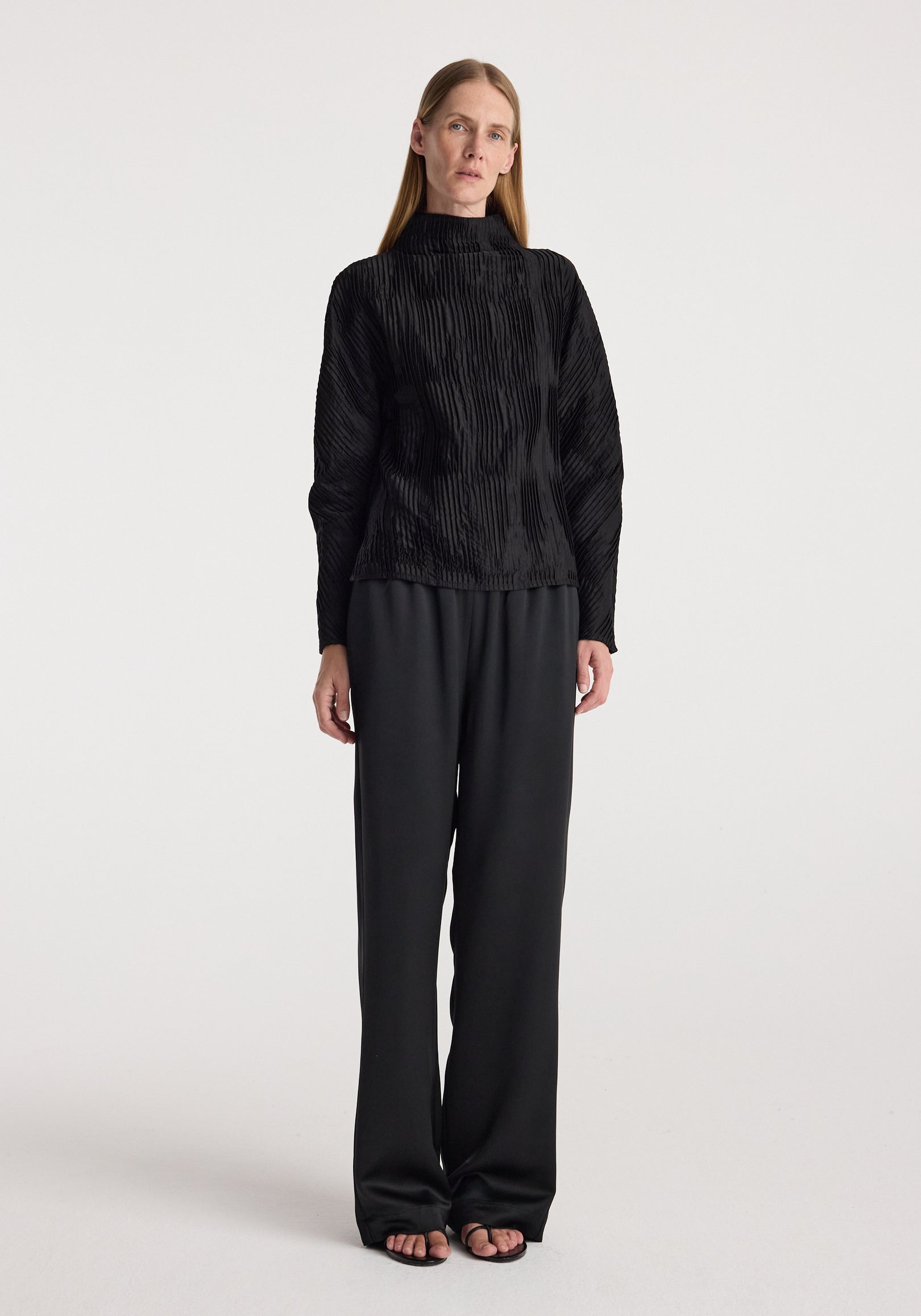 Woman wearing relaxed fit satin trousers | black paired with a textured black long-sleeve top, standing against a plain white background.
