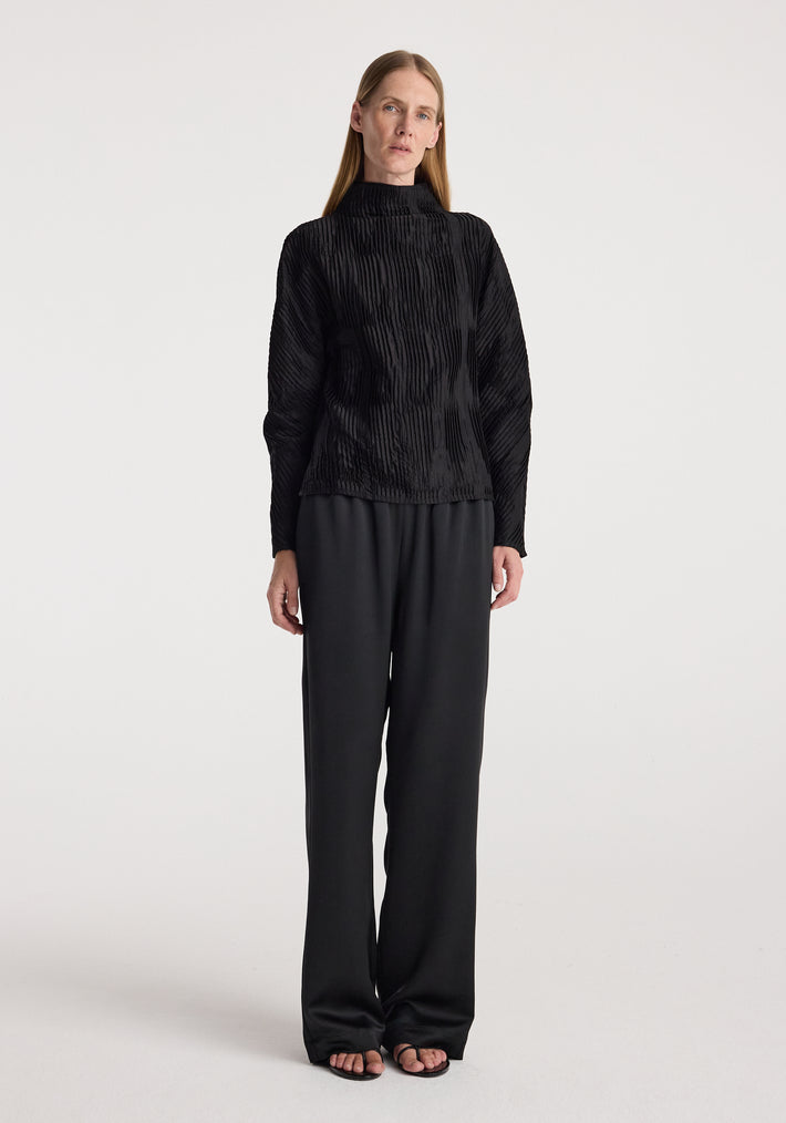 Woman wearing relaxed fit satin trousers | black paired with a textured black long-sleeve top, standing against a plain white background.