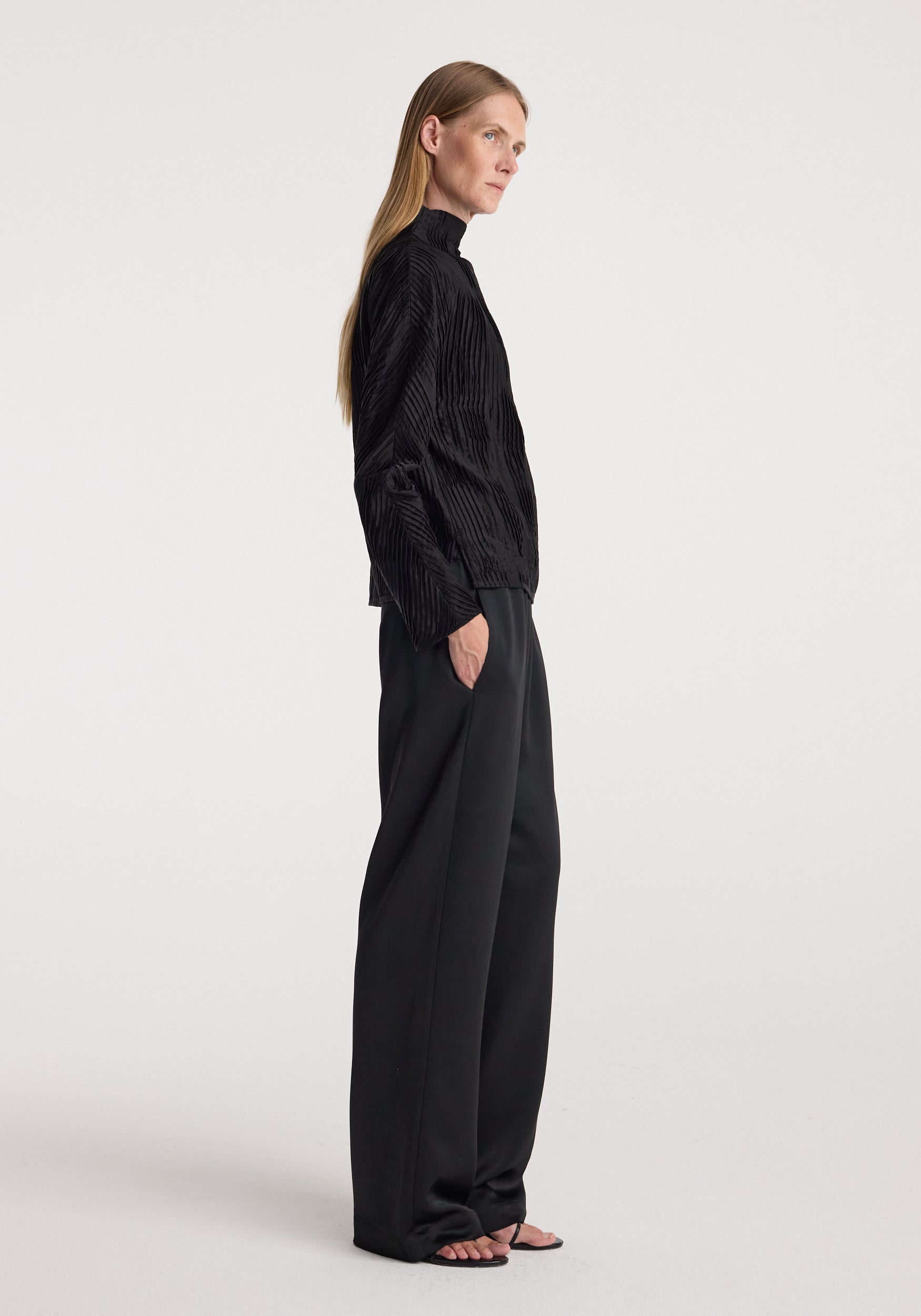 Woman standing in profile wearing relaxed fit satin trousers | black paired with a long-sleeve black textured top and open-toe sandals against a plain background.