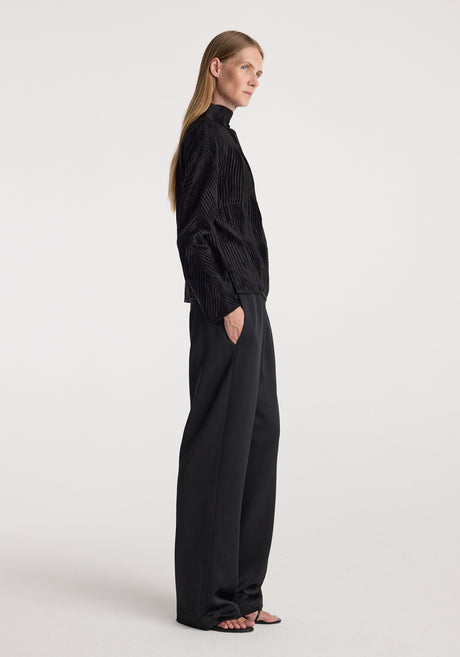 Woman wearing relaxed fit satin trousers | black paired with a textured black long-sleeve top, standing against a plain white background.