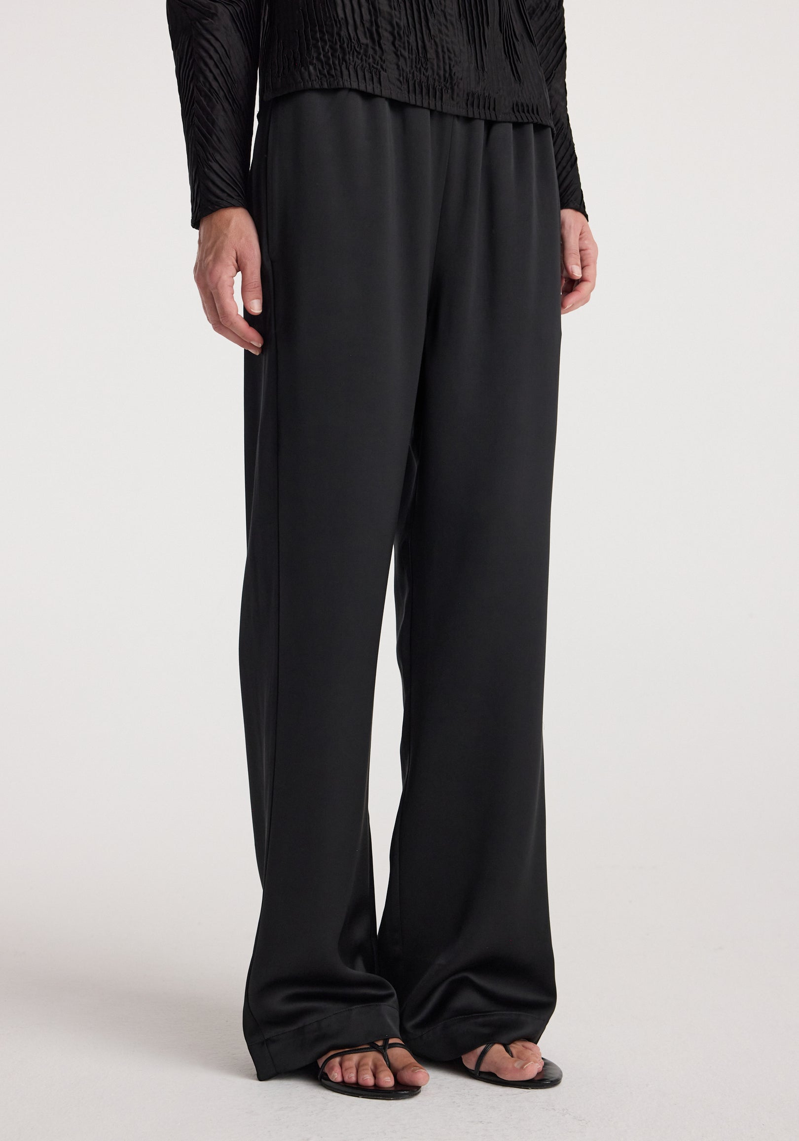 Front view of a person wearing relaxed fit satin trousers | black paired with a black textured long-sleeve top and black sandals against a plain background.