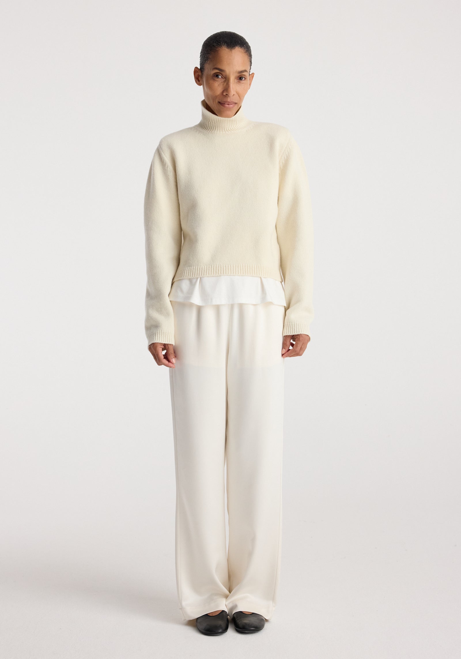Woman wearing cream-colored relaxed fit satin trousers paired with a light sweater and black shoes against a plain white background, showcasing Relaxed fit satin trousers | cream.