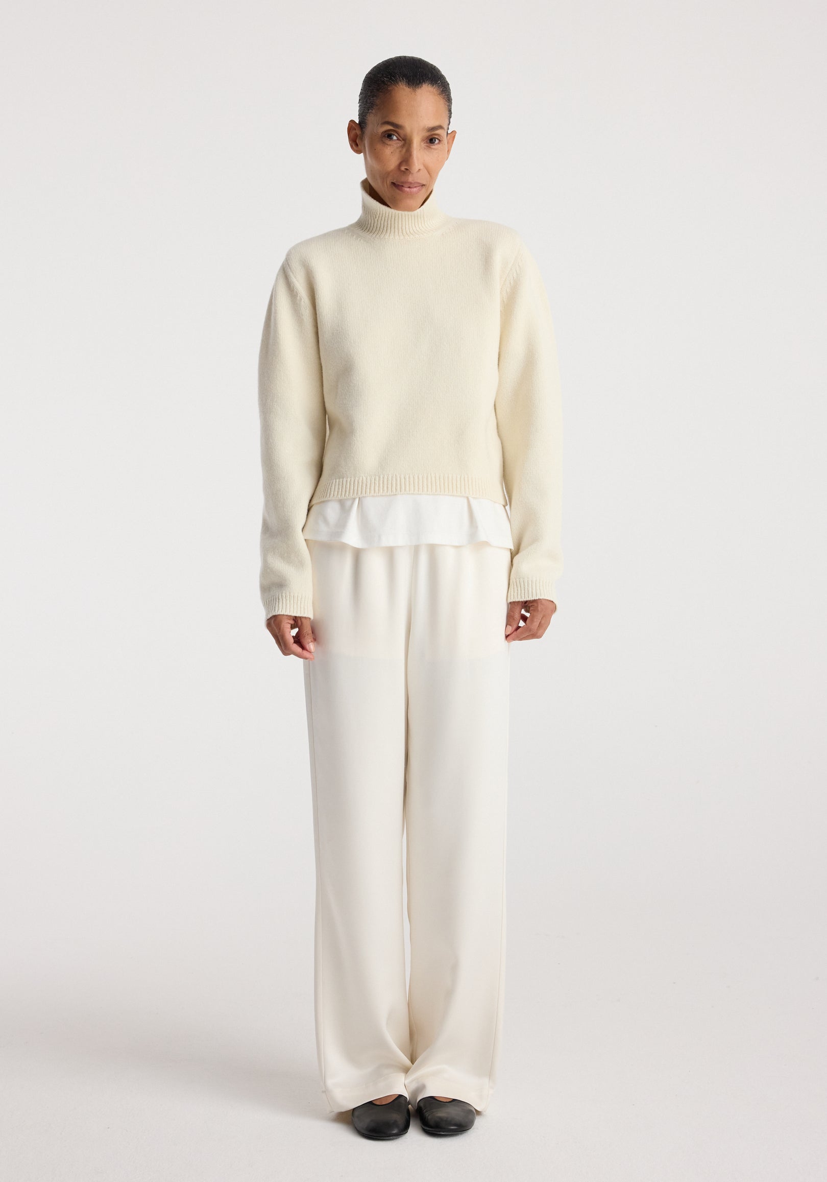 Woman wearing cream-colored relaxed fit satin trousers paired with a light sweater and black shoes against a plain white background, showcasing Relaxed fit satin trousers | cream.