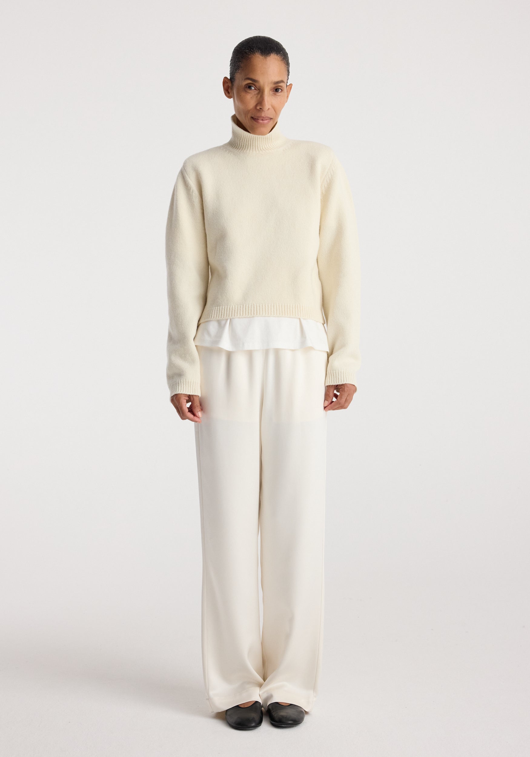 Woman wearing cream-colored relaxed fit satin trousers paired with a light sweater and black shoes against a plain white background, showcasing Relaxed fit satin trousers | cream.