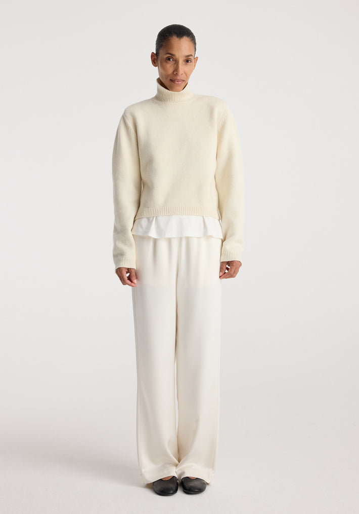 Woman wearing cream-colored relaxed fit satin trousers paired with a light sweater and black shoes against a plain white background, showcasing Relaxed fit satin trousers | cream.