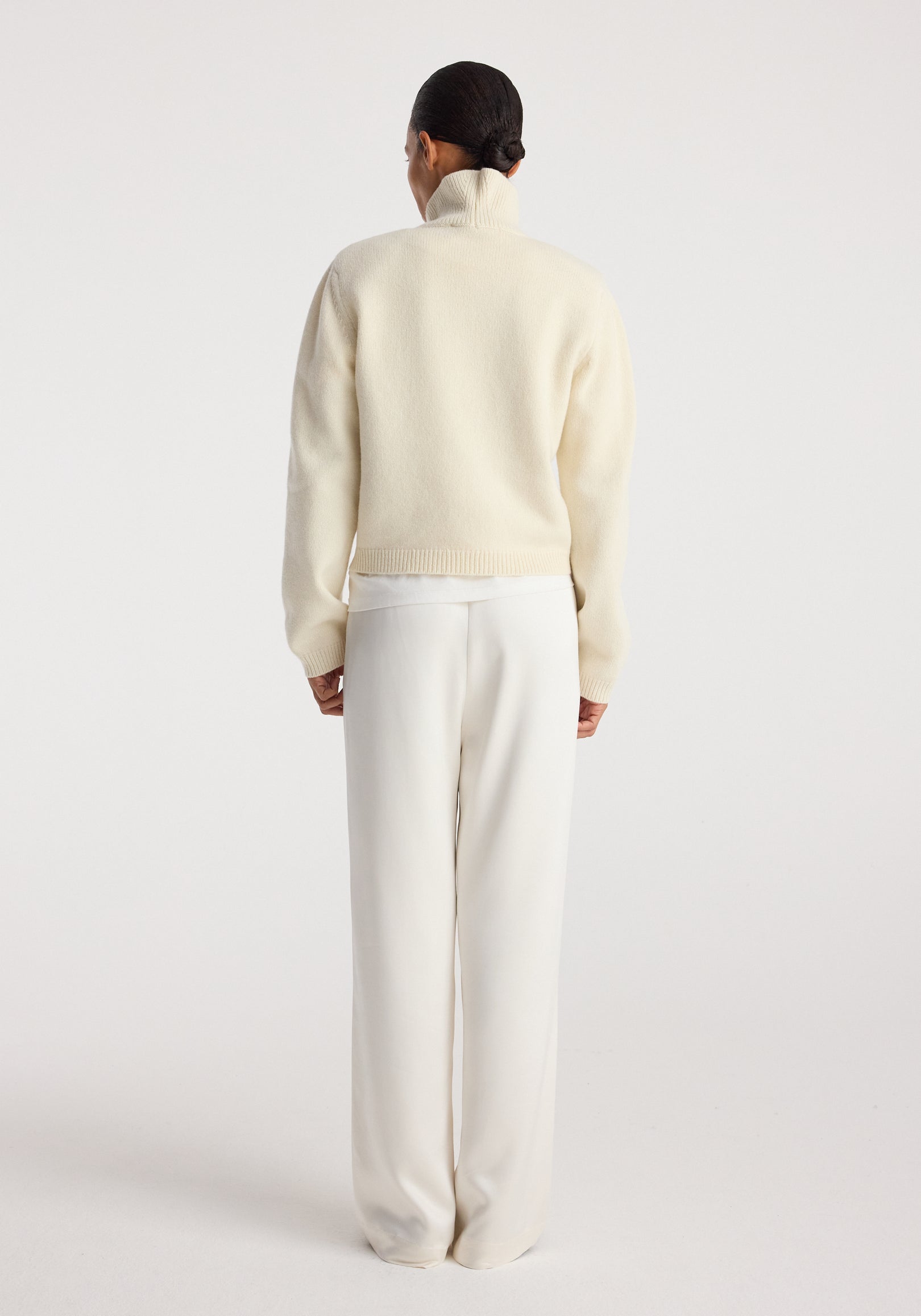 Woman wearing cream-colored relaxed fit satin trousers standing with her back to the camera against a plain background, paired with a light-colored sweater, showcasing the smooth texture of the Relaxed fit satin trousers | cream.