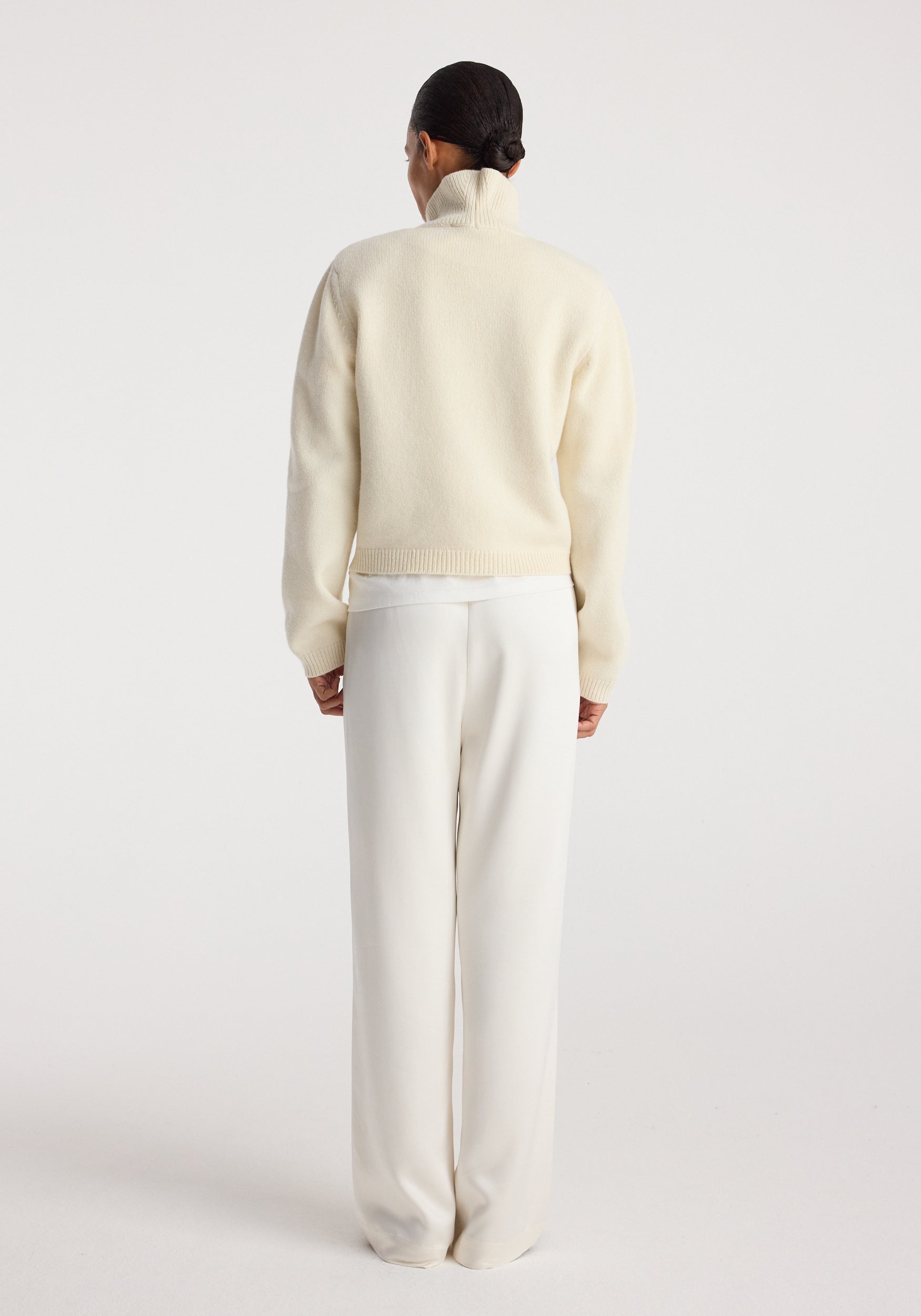 Woman wearing cream-colored relaxed fit satin trousers standing with her back to the camera against a plain background, paired with a light-colored sweater, showcasing the smooth texture of the Relaxed fit satin trousers | cream.
