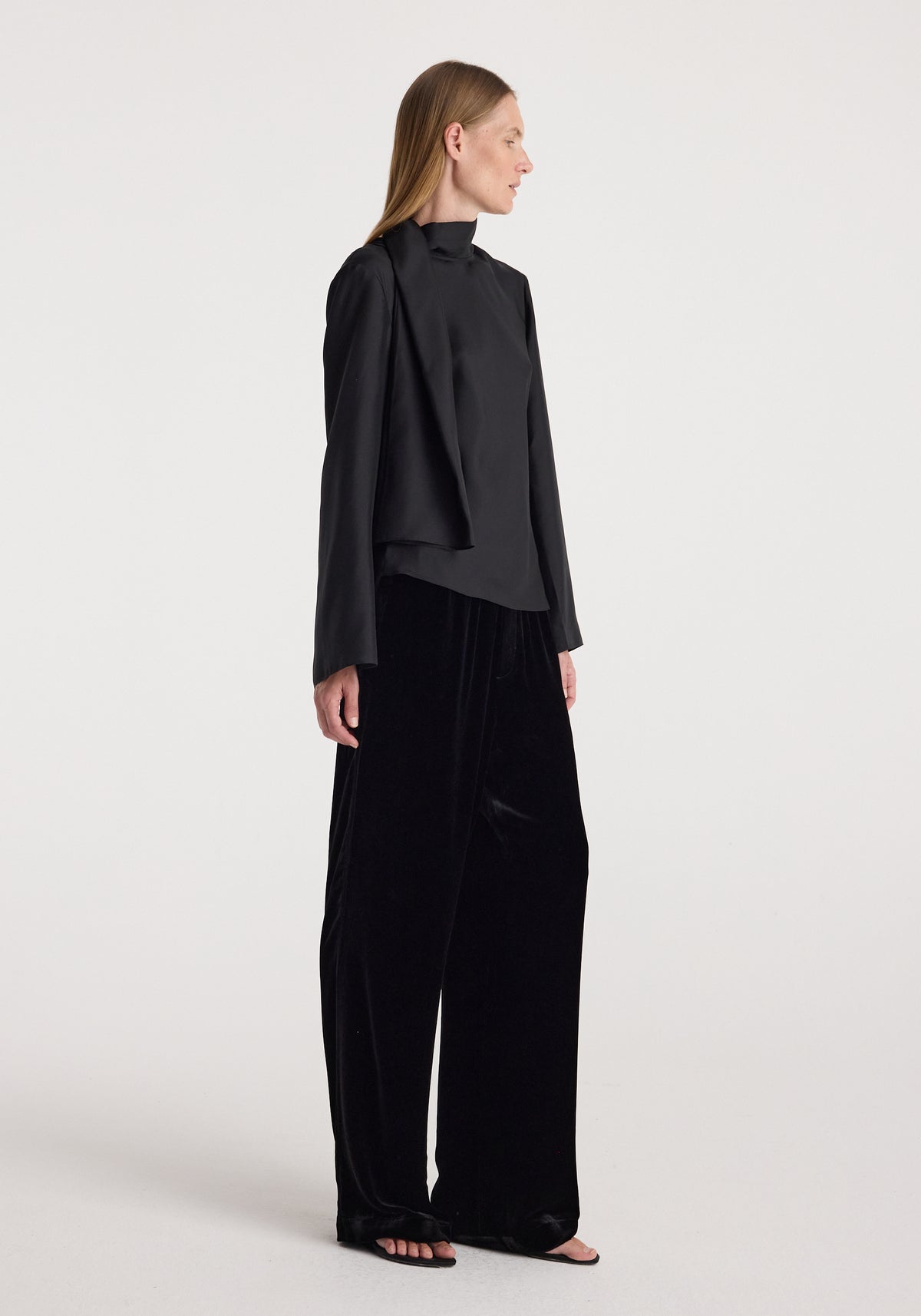 A woman wearing black, loose-fitting velvet relaxed trousers | black paired with a long-sleeve black top, standing in profile against a plain white background.