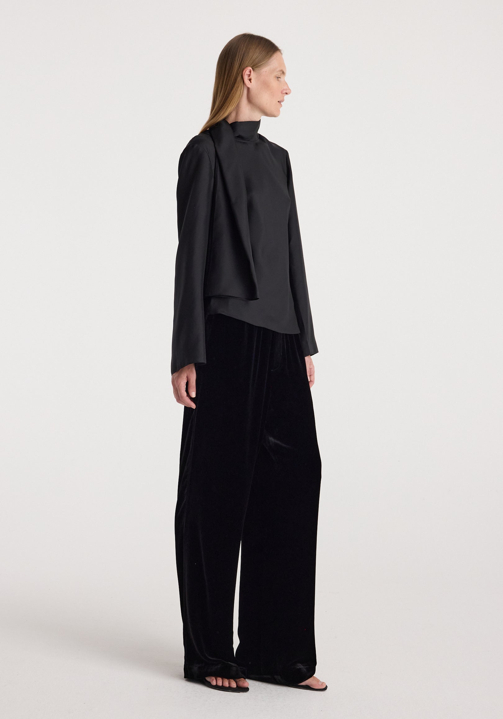A woman wearing black, loose-fitting velvet relaxed trousers | black paired with a long-sleeve black top, standing in profile against a plain white background.
