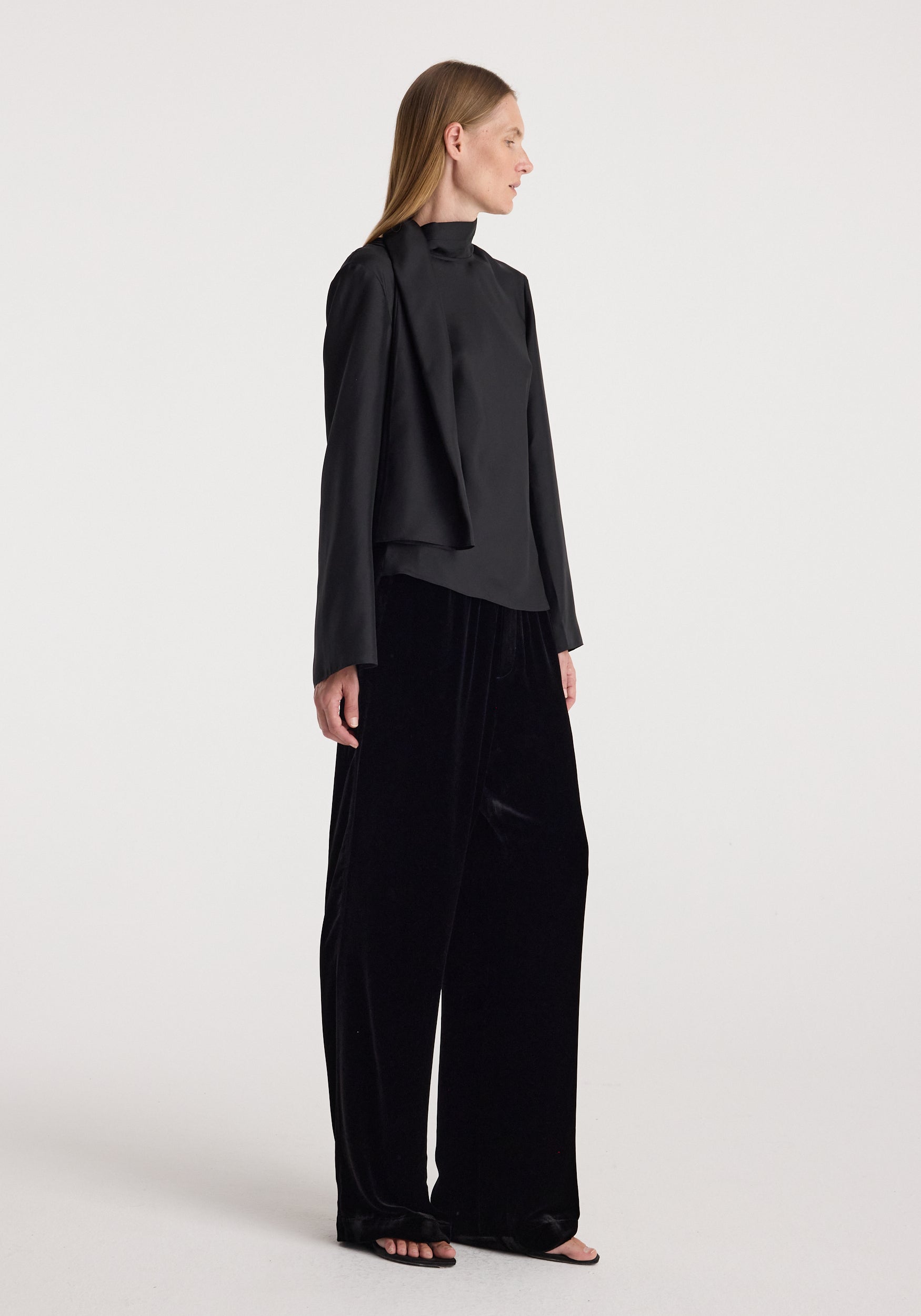 A woman wearing black, loose-fitting velvet relaxed trousers | black paired with a long-sleeve black top, standing in profile against a plain white background.