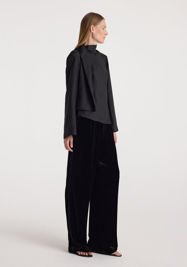 A woman wearing black, loose-fitting velvet relaxed trousers | black paired with a long-sleeve black top, standing in profile against a plain white background.