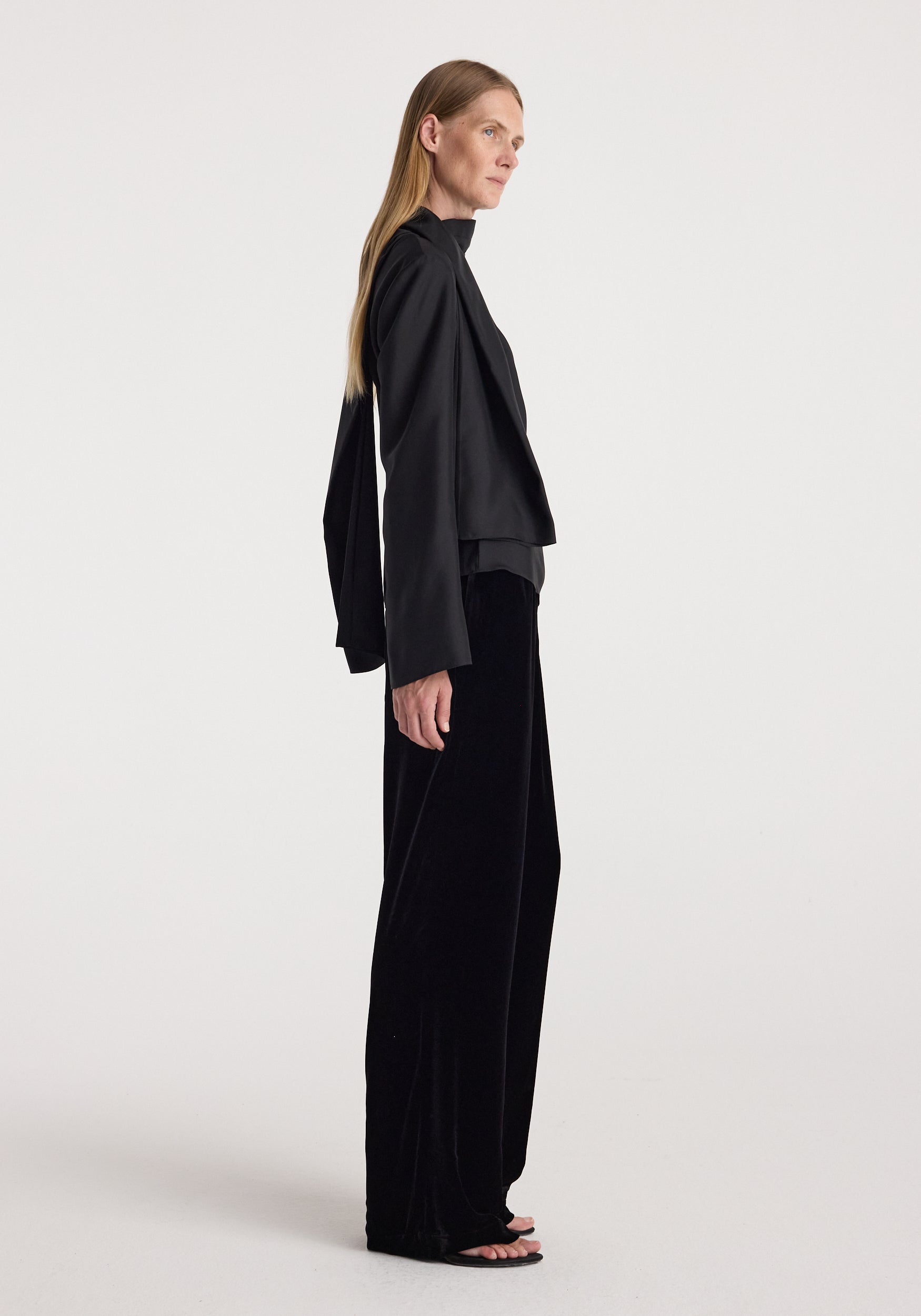 Side view of a woman wearing Velvet relaxed trousers | black with a loose fit, paired with a long-sleeve black top, standing against a plain light background.