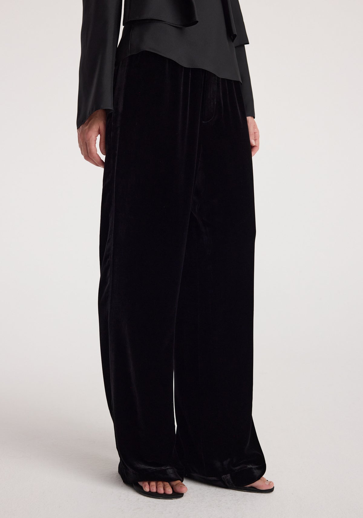 Close-up of a person wearing black velvet relaxed trousers | black with a loose fit and smooth texture, paired with black open-toe sandals and a black top.