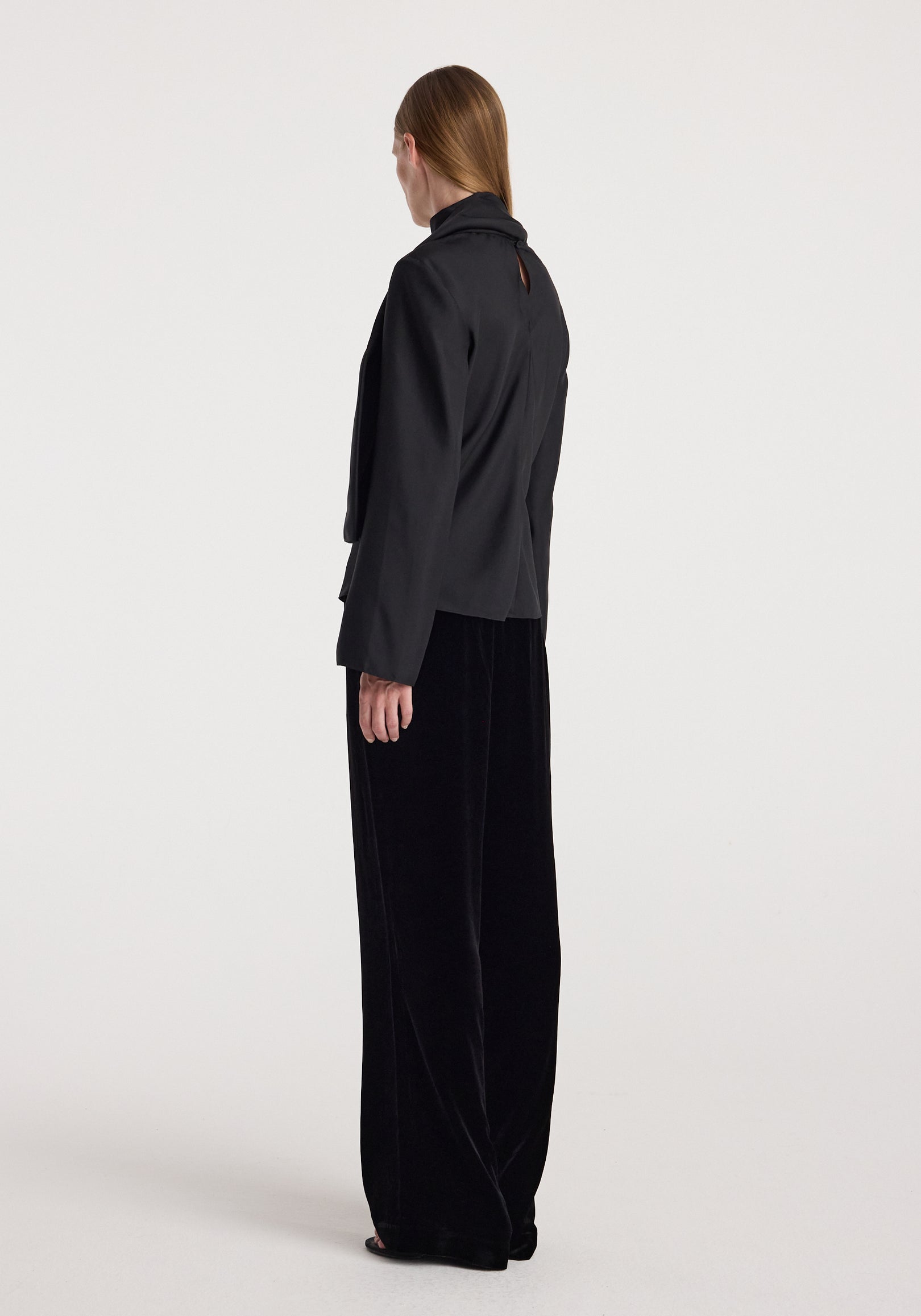 Woman wearing black velvet relaxed trousers | black with a loose fit and wide legs, paired with a black long-sleeve top, standing against a plain light background.
