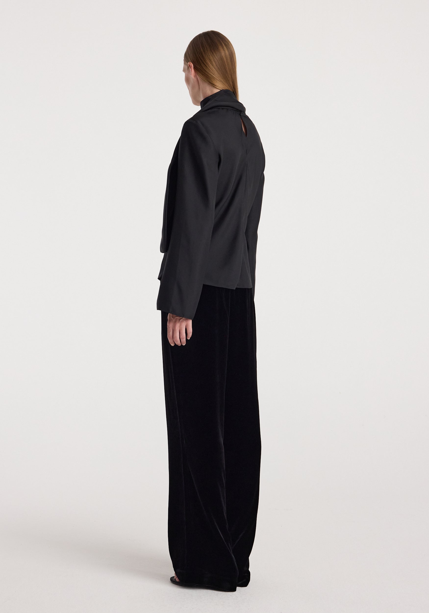Woman wearing black velvet relaxed trousers | black with a loose fit and wide legs, paired with a black long-sleeve top, standing against a plain light background.