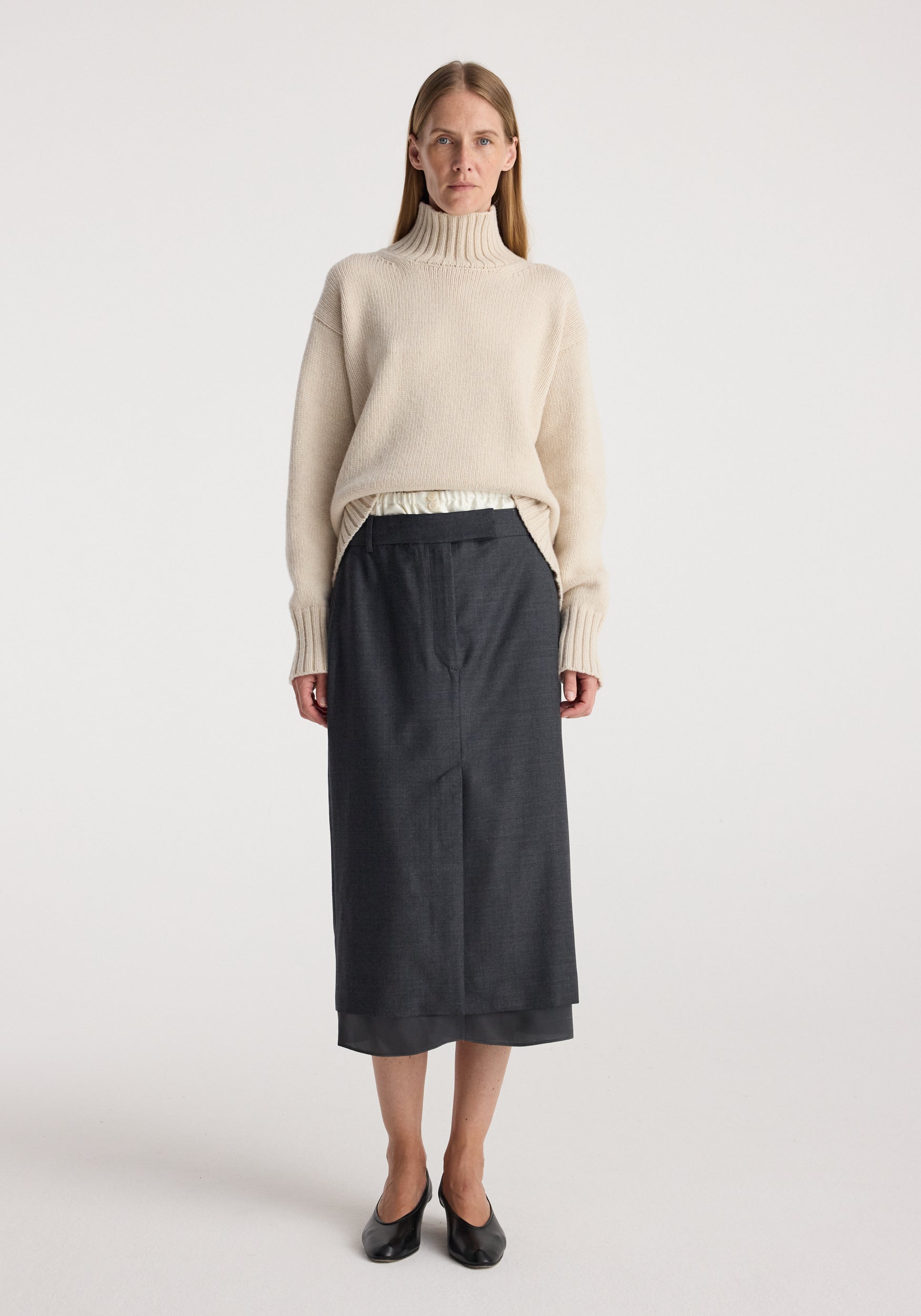 Woman wearing a light beige turtleneck sweater paired with a dark grey melange Boxer detailed skirt | dark grey melange, standing against a plain white background.
