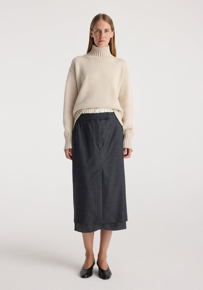 Woman wearing a light beige turtleneck sweater paired with a dark grey melange Boxer detailed skirt | dark grey melange, standing against a plain white background.