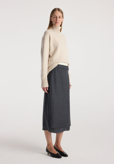 Woman wearing a light beige turtleneck sweater paired with a dark grey melange Boxer detailed skirt | dark grey melange, standing against a plain white background.