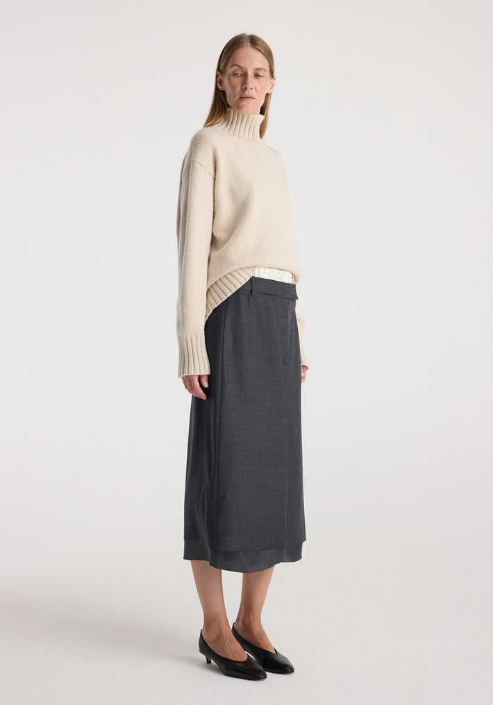 Woman wearing a beige turtleneck sweater paired with a Boxer detailed skirt | dark grey melange, styled with black low-heeled shoes in a minimalist setting.